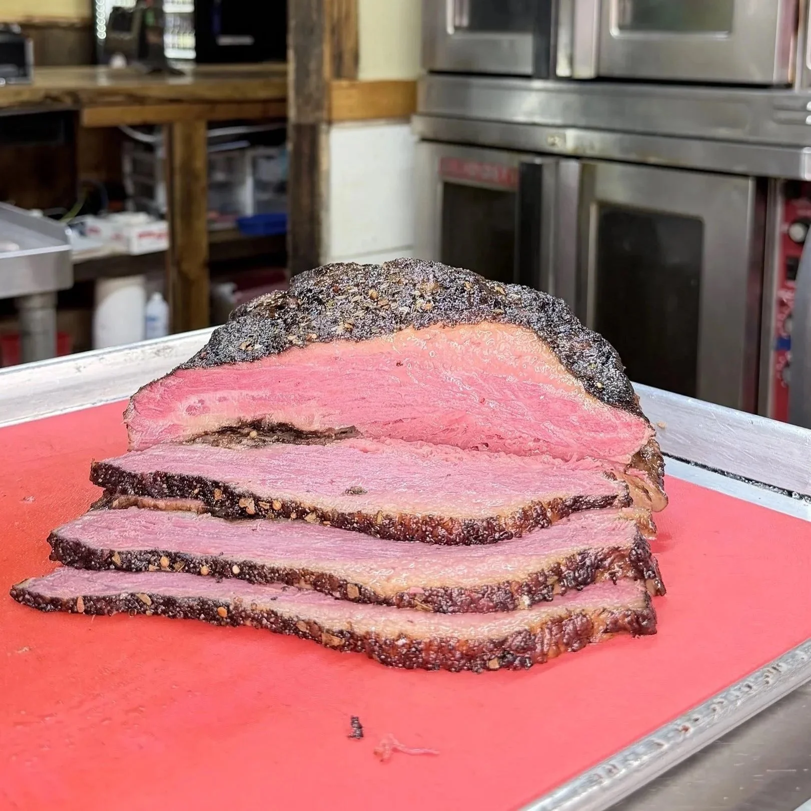 Slices of rare smoked beef rubbed with black pepper on a pink cutting board with a commercial kitchen background.