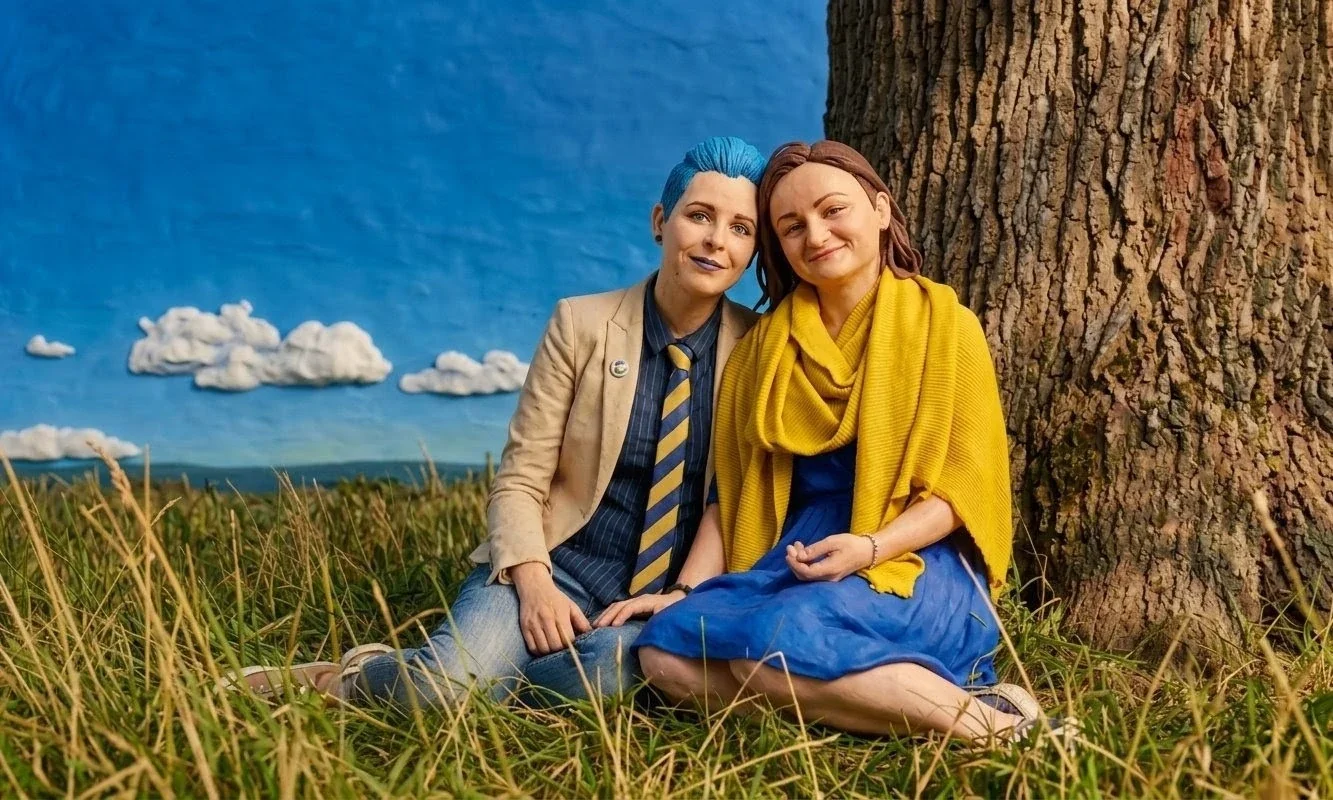 Two women sitting on grass next to a large tree, smiling at the camera with sky and clouds in the background.