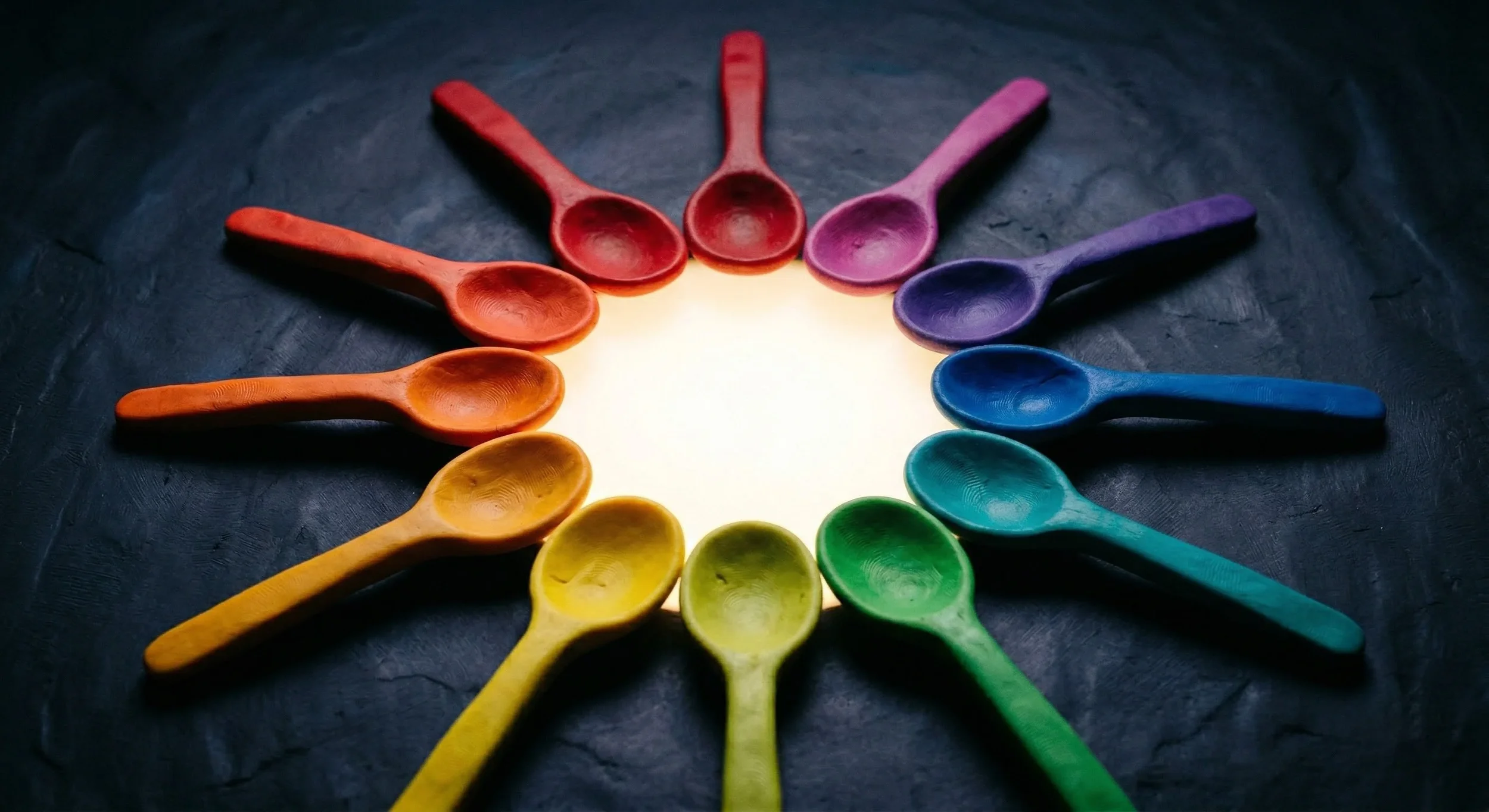 Colorful wooden spoons arranged in a circle on a dark surface, illuminated from the center.