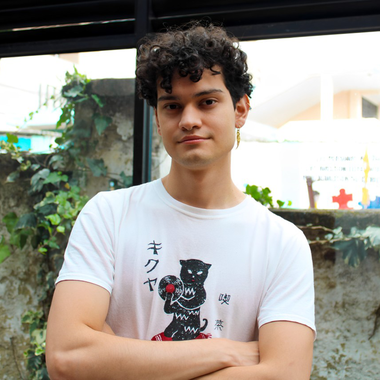 A young man with curly dark hair, wearing a white T-shirt with a graphic of a black cat holding a red ball and Japanese characters, standing with arms crossed outdoors.