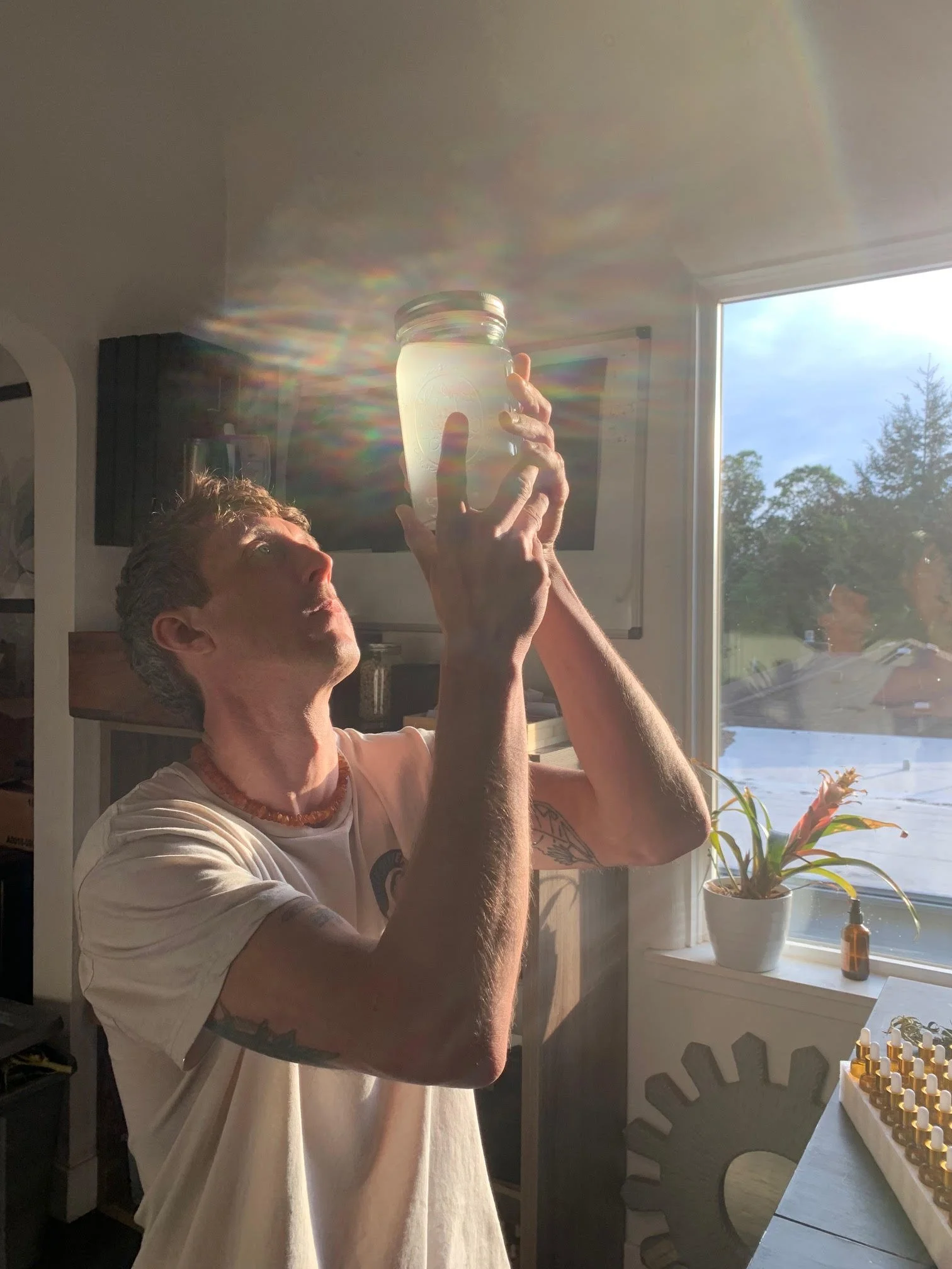 Person holding a jar with sunlight creating rainbow reflections on the ceiling, near a window with a plant and various bottles.