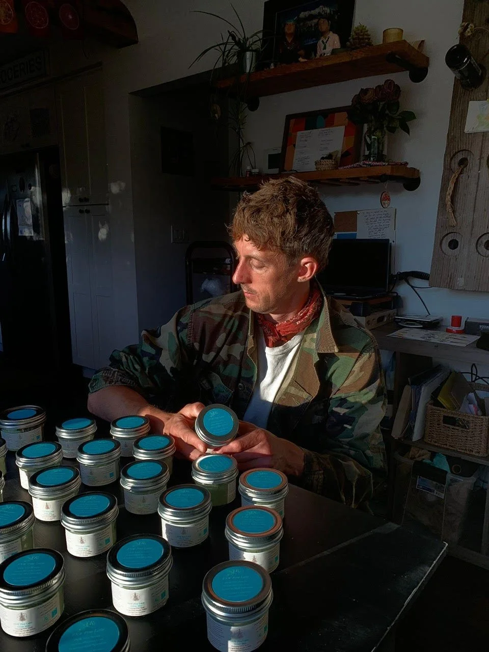 A man in camouflage jacket and red neckerchief examining small jars with blue labels on a table inside a room.