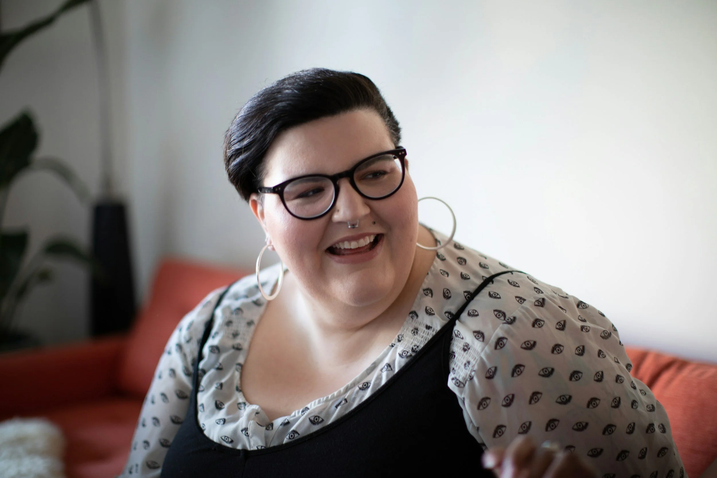 Smiling person with glasses, nose and upper lip piercing wearing a white shirt with eyes on it andt in herapy for highly sensitive person Los Angeles