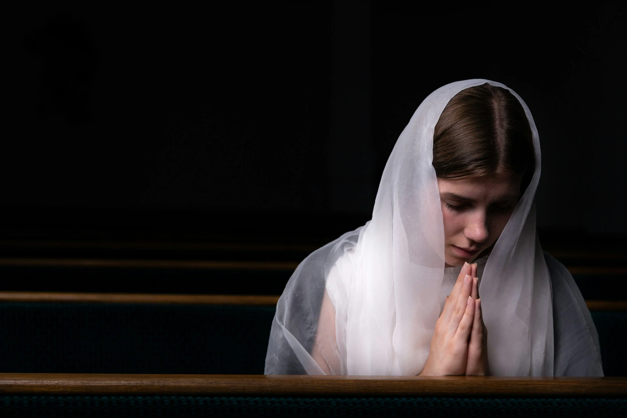 Person seated in a church with head bowed and hands together, representing the emotional impact of religious trauma, spiritual harm, and institutional abandonment.