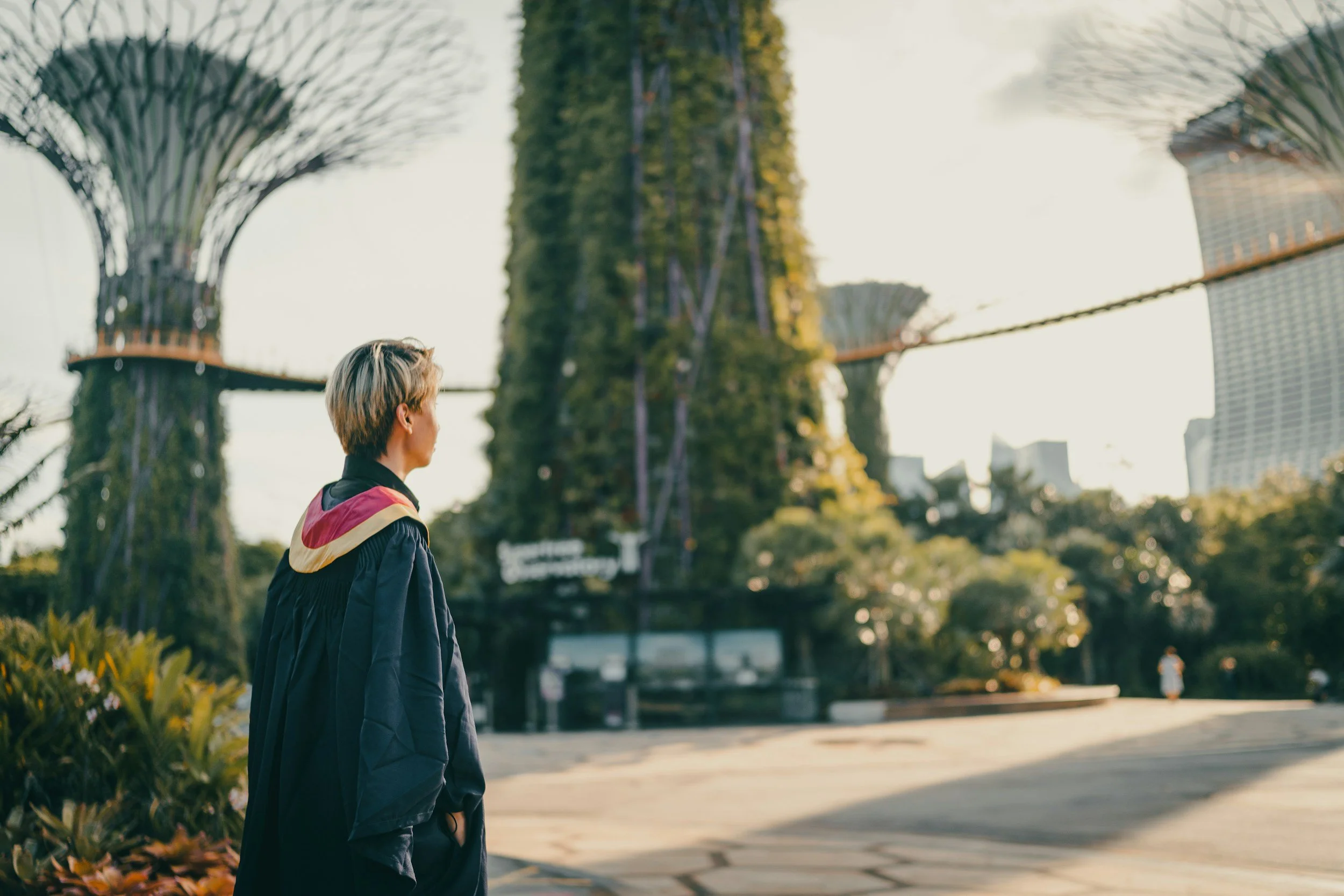 A graduate in academic regalia standing alone in a campus landscape, reflecting on the emotional impact of grad school and art school trauma.