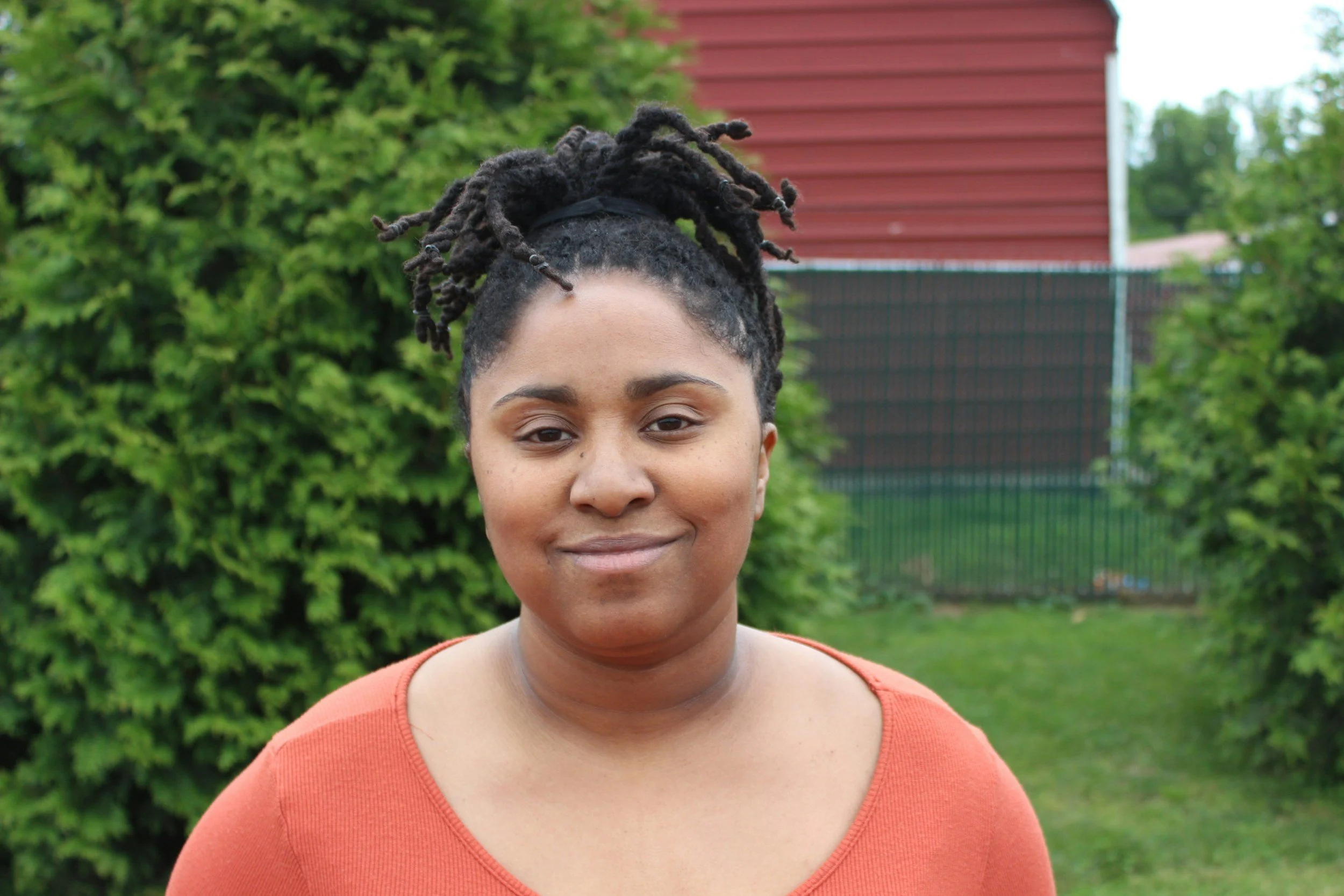 A person with dark hair in a high bun of locs smiling gently at the camera in a backyard setting with green bushes.