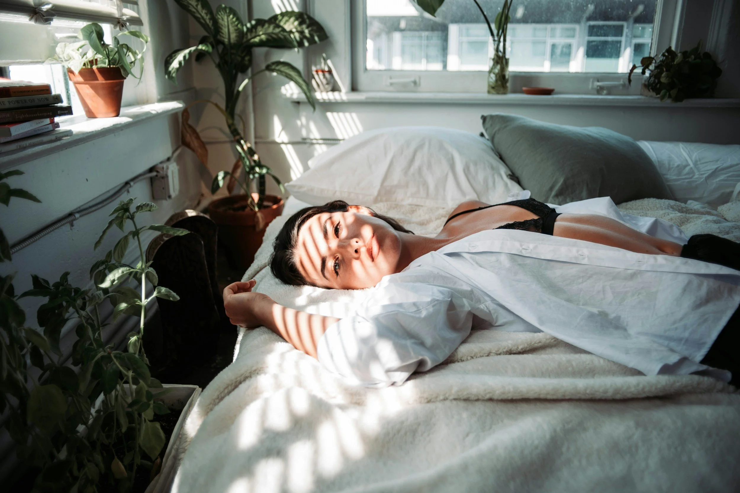 Person lying in bed in soft window light surrounded by houseplants, suggesting rest and nervous system recovery.