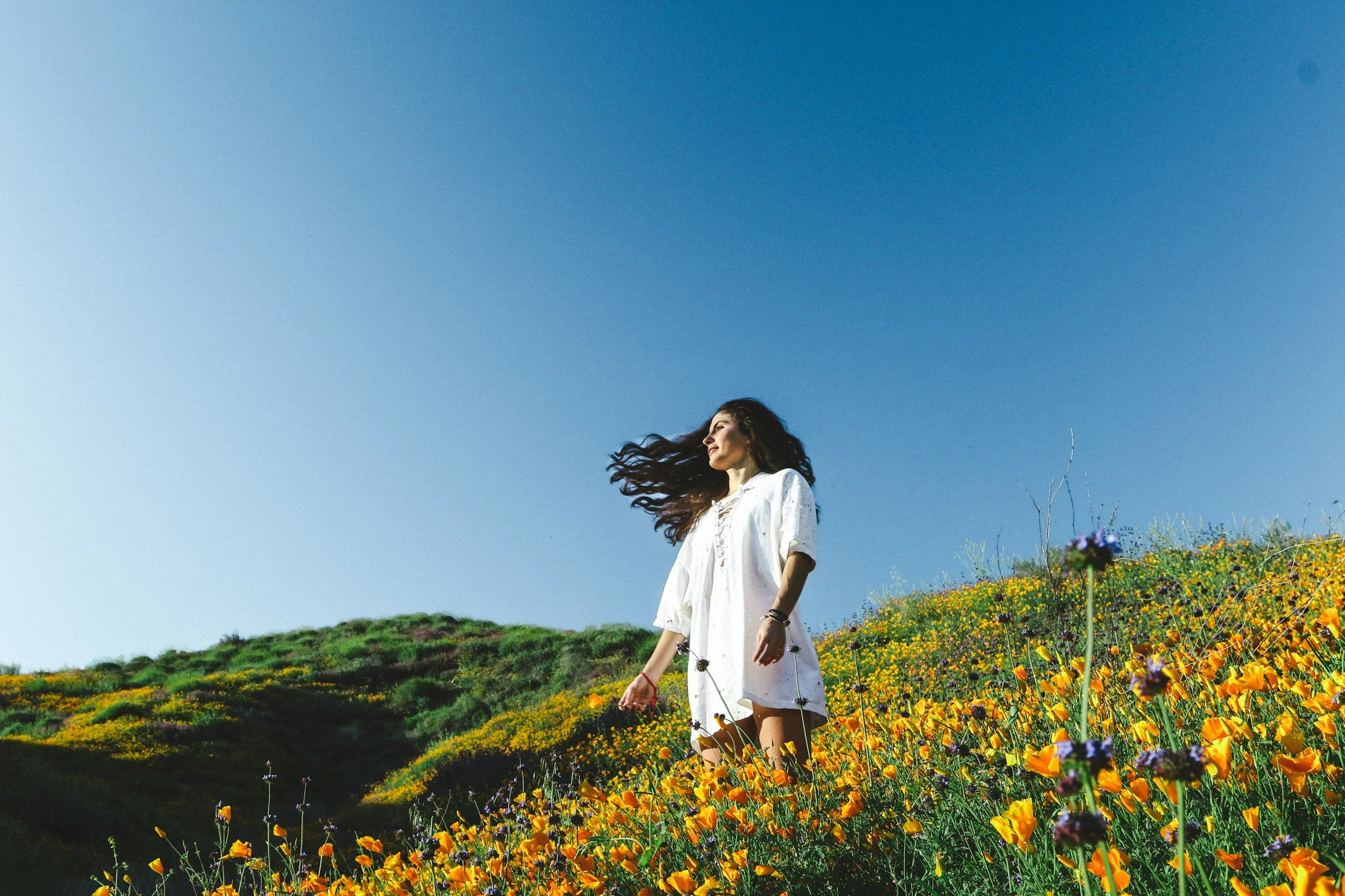 Person in a white dress walking through a field of wildflowers beneath a wide blue sky.