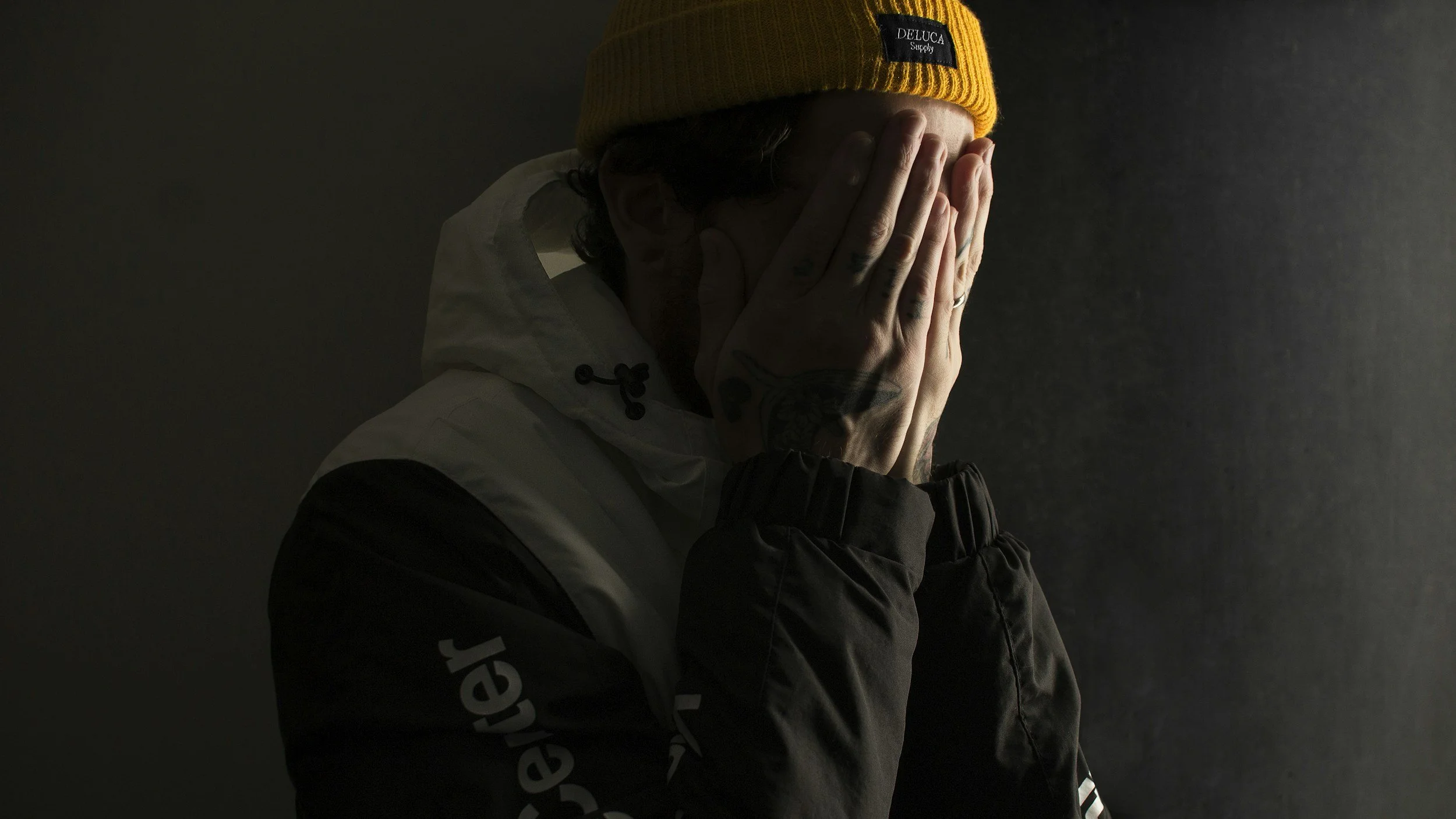 Person in a yellow beanie covering their face with tattooed hands in a dark, shadowed room, illustrating how high-control environments feel from the inside—overwhelmed, hidden, and ashamed.