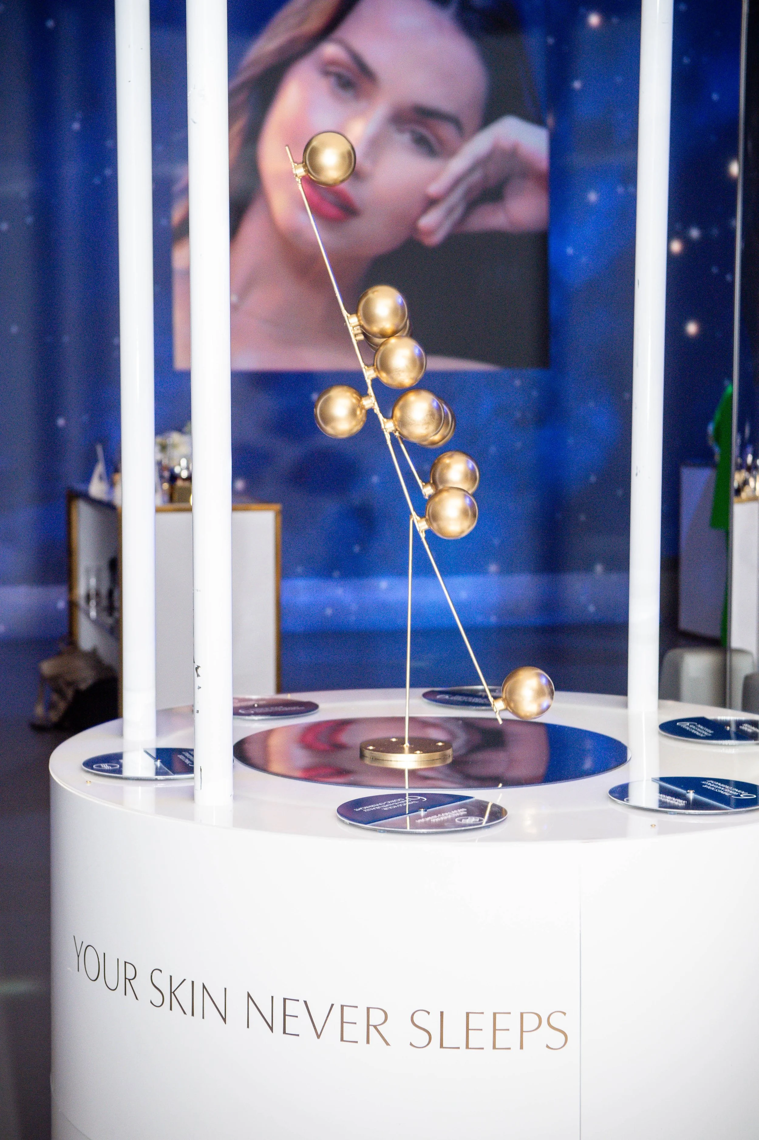 Display with a golden sculpture of spheres on wires, set on a white circular table with the phrase "Your skin never sleeps." In the background, a large poster of a woman's face with makeup and a soft expression.
