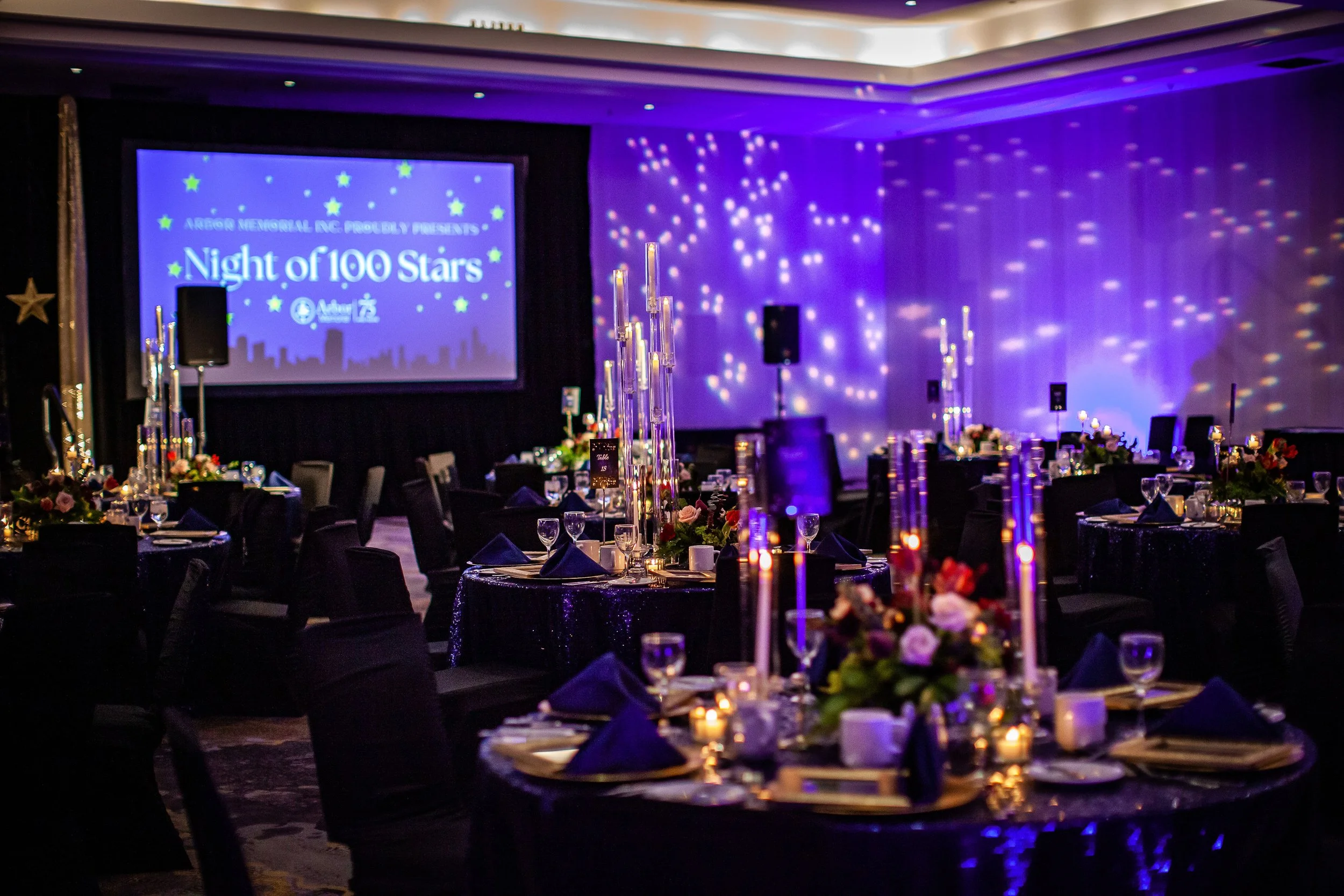 Ballroom decorated for a formal event with round tables, dark tablecloths, floral centerpieces, tall candles, and purple lighting. A large screen displays 'Night of 100 Stars' with a city skyline graphic in the background.
