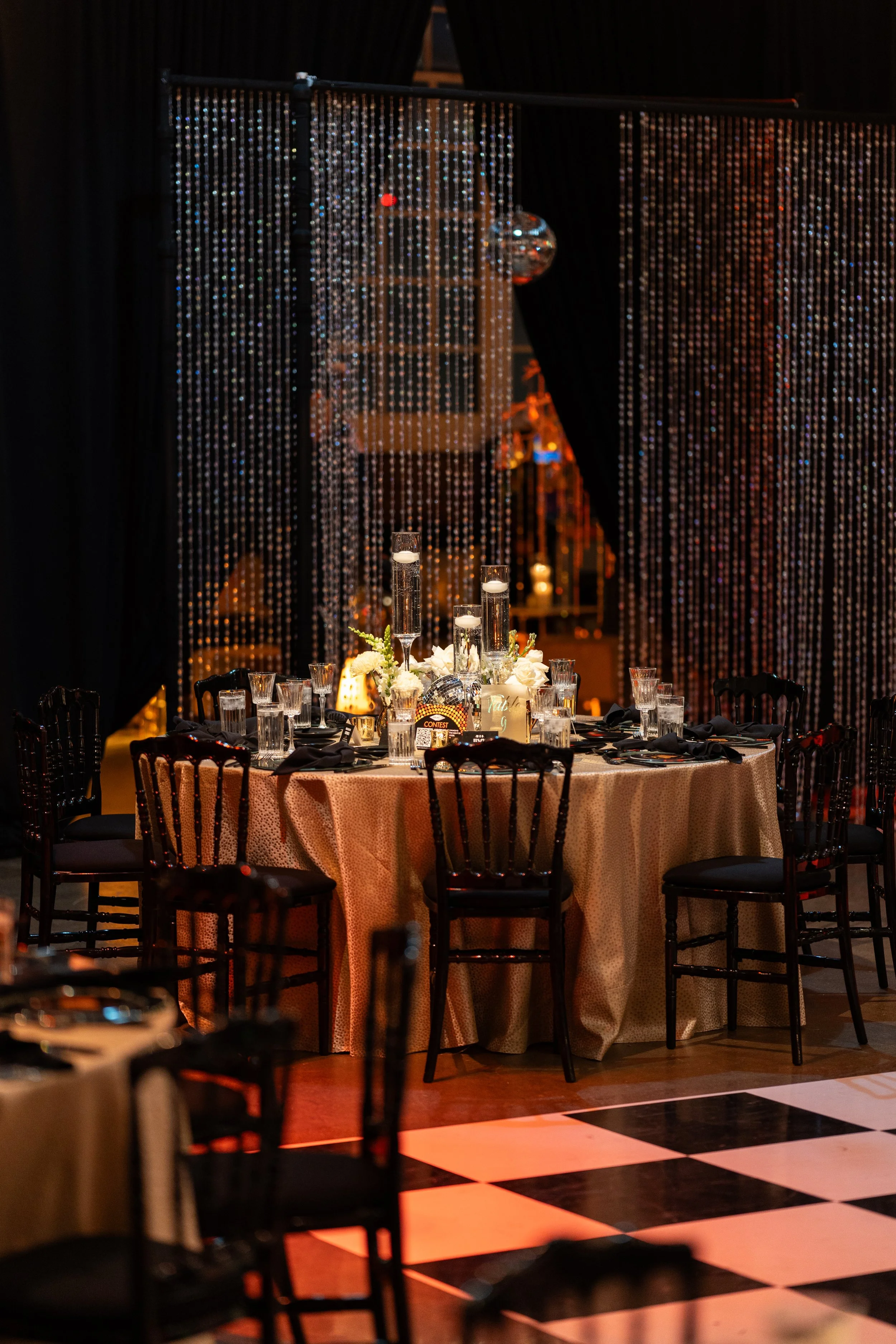 Decorated banquet table with candles and flowers, set for an event, with beaded curtains and disco ball in the background.