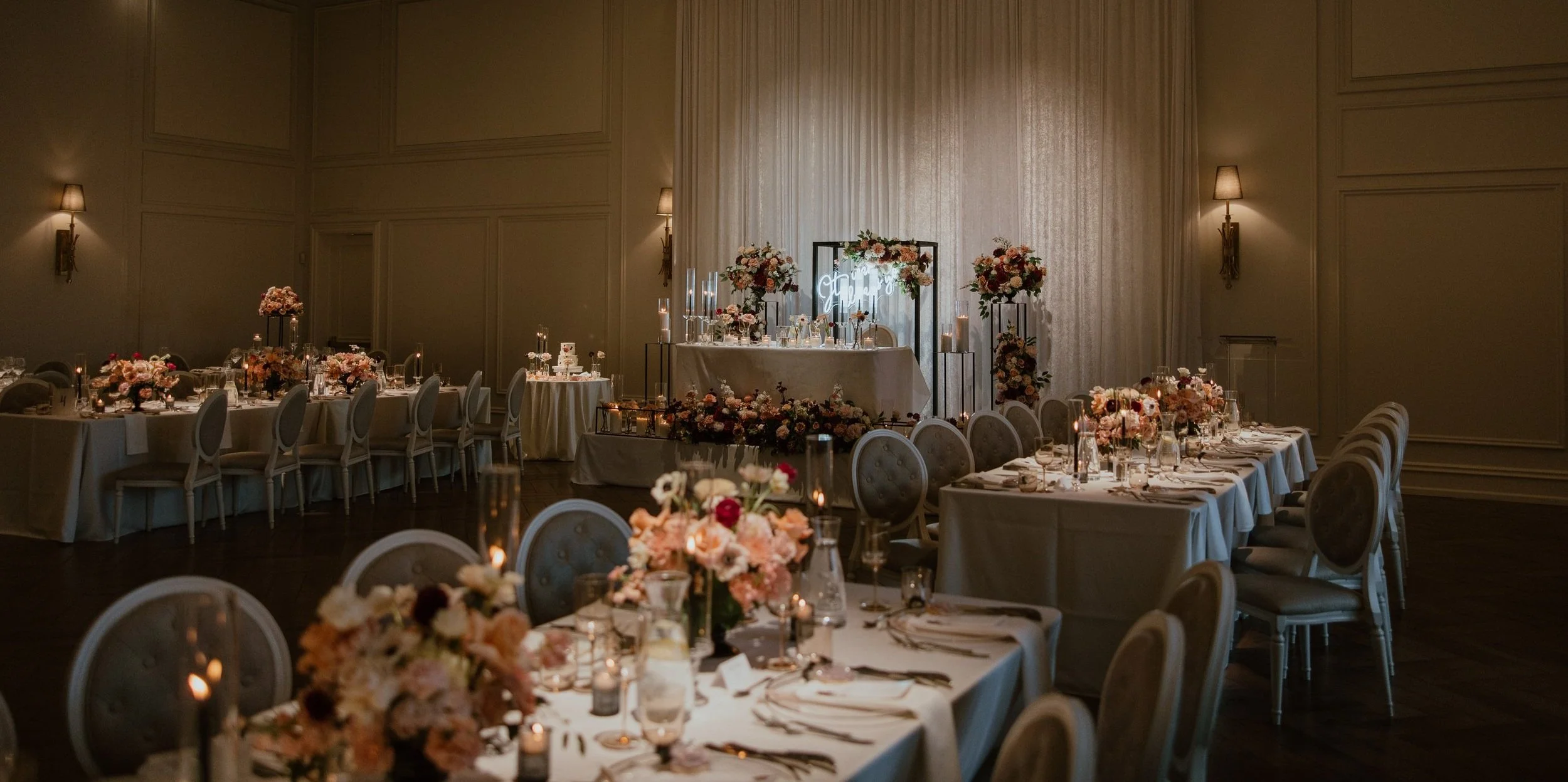 Wedding reception hall decorated with pink and white flowers, candles, and elegant table settings, with a sweetheart table and a neon sign in the background.