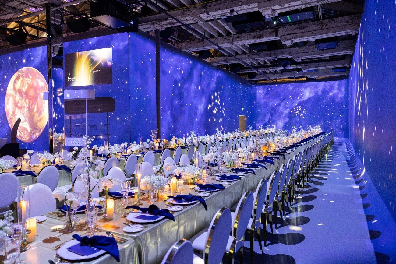 Elegant banquet hall with long dining tables set with white linens, navy blue napkins, flower arrangements, and candles, illuminated by starry blue galaxy-themed walls and ceiling.