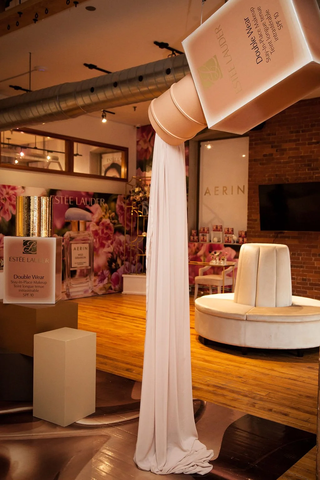 Interior of a beauty or skincare store or studio with a large beauty product display, a white draped fabric, and seating area with a white chair. Promotional posters and products featuring Estée Lauder and Aerin are visible.