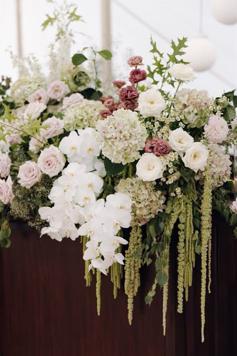 A floral arrangement with white orchids, pink roses, hydrangeas, and hanging greenery, placed on a dark wooden surface.
