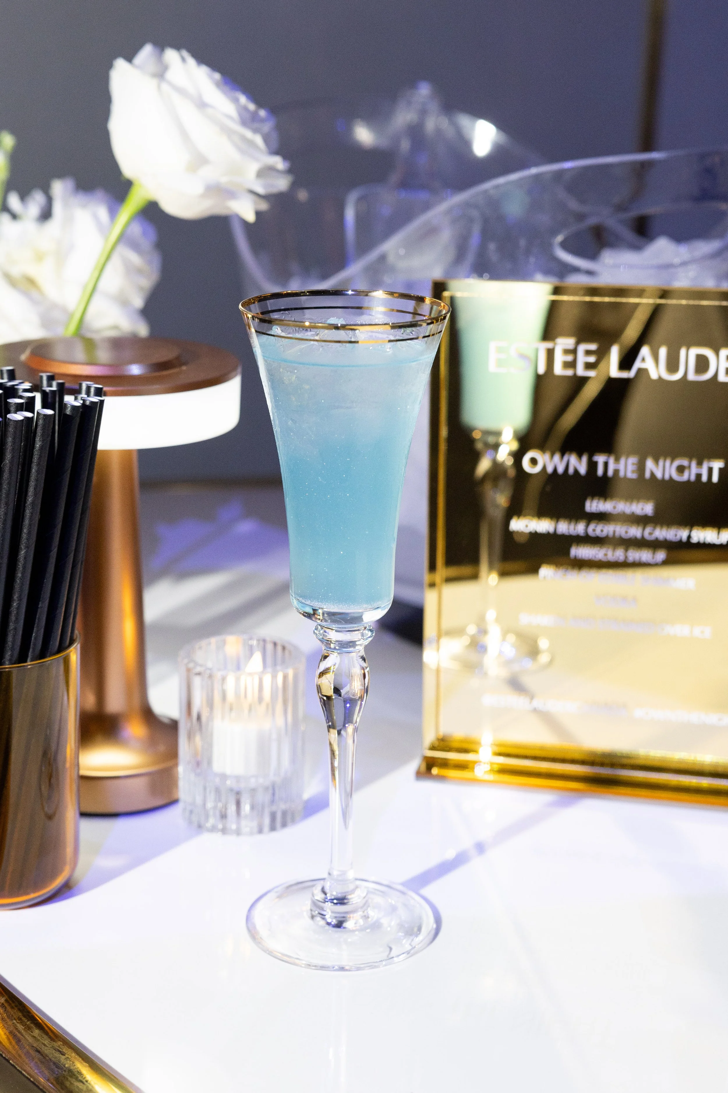 A light blue cocktail in a tall, elegant glass with a gold rim, placed on a white surface with decorative items and a black display in the background.