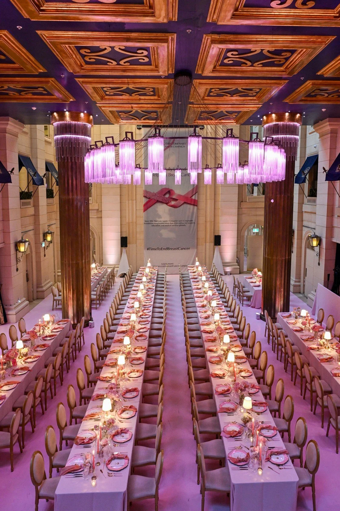 A large, elegant banquet hall decorated for a breast cancer awareness event, with long tables set with pink napkins, flowers, and small lamps, and a prominent banner with a pink ribbon symbol in the background.