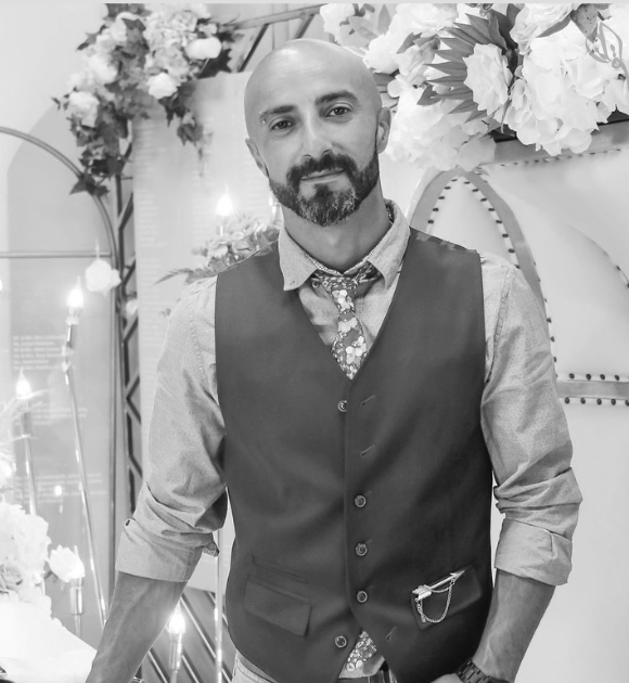 A man with a beard and shaved head wearing a vest, shirt, and patterned tie in a decorated setting with flowers and lights.