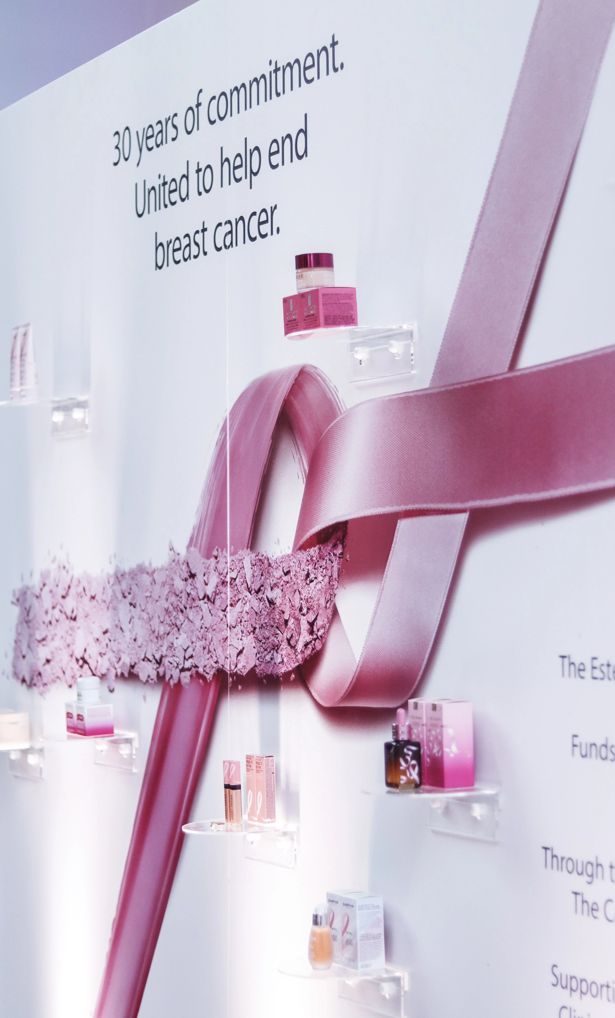 Display of pink-themed skincare or cosmetic products arranged on clear shelves with a pink ribbon and crushed pink powder underneath. A large white wall features black text celebrating 30 years of commitment to helping end breast cancer.