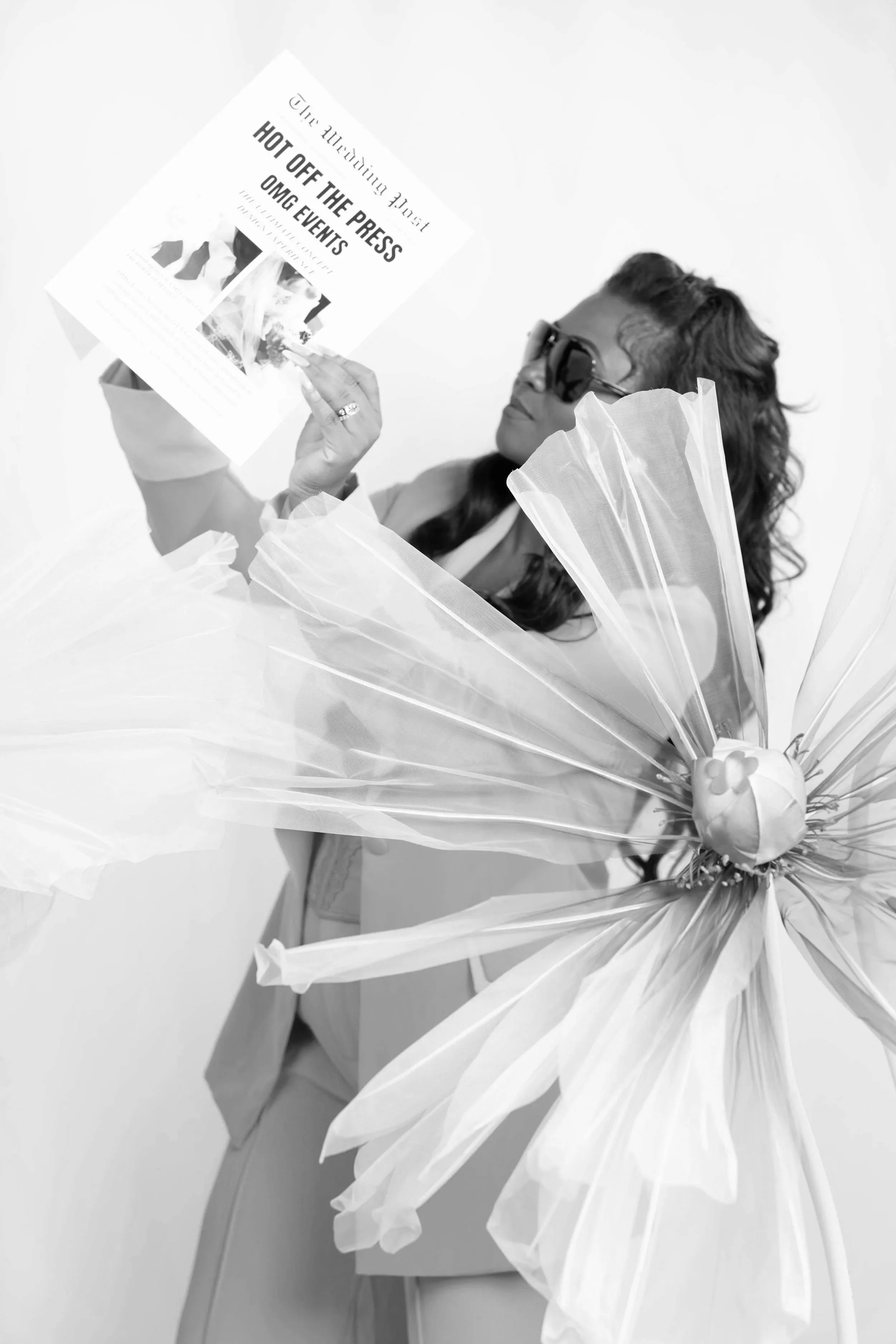 A woman wearing sunglasses and a jacket, holding a newspaper and standing beside a large decorative flower made of paper or fabric.