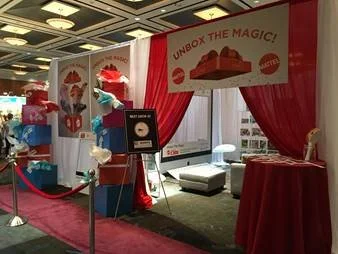 Trade show booth with red curtains, a banner that says 'Unbox the Magic!', and a chocolate cake display.