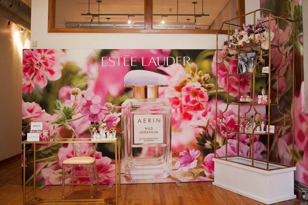 Display booth for Estée Lauder's AERIN Wild Geranium perfume featuring a large floral backdrop, a big perfume bottle image, a small table with a chair, and a gold shelf with perfume bottles and beauty products.