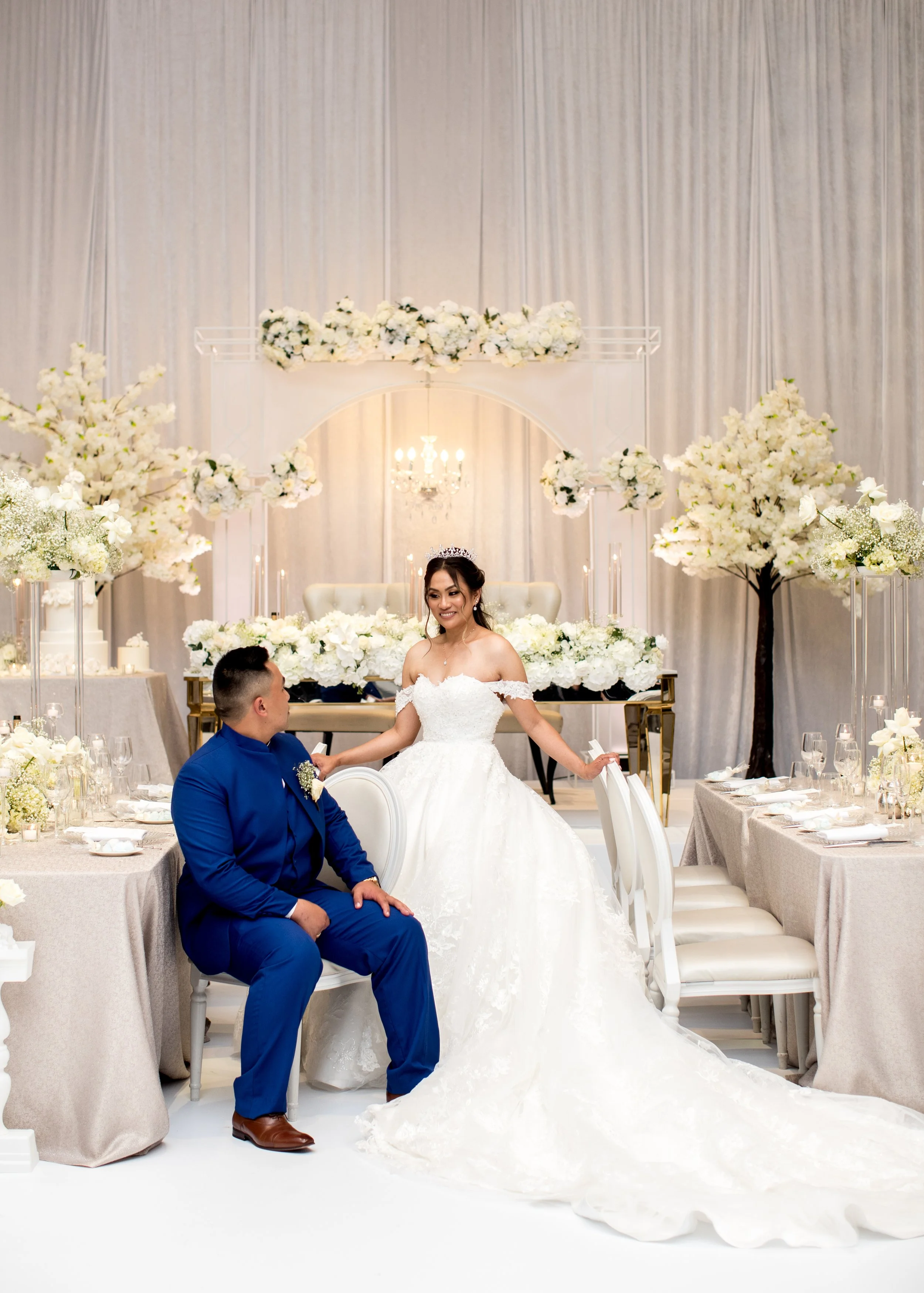 A bride in a white wedding dress and tiara sitting on a chair, and a groom in a blue suit sitting beside her.