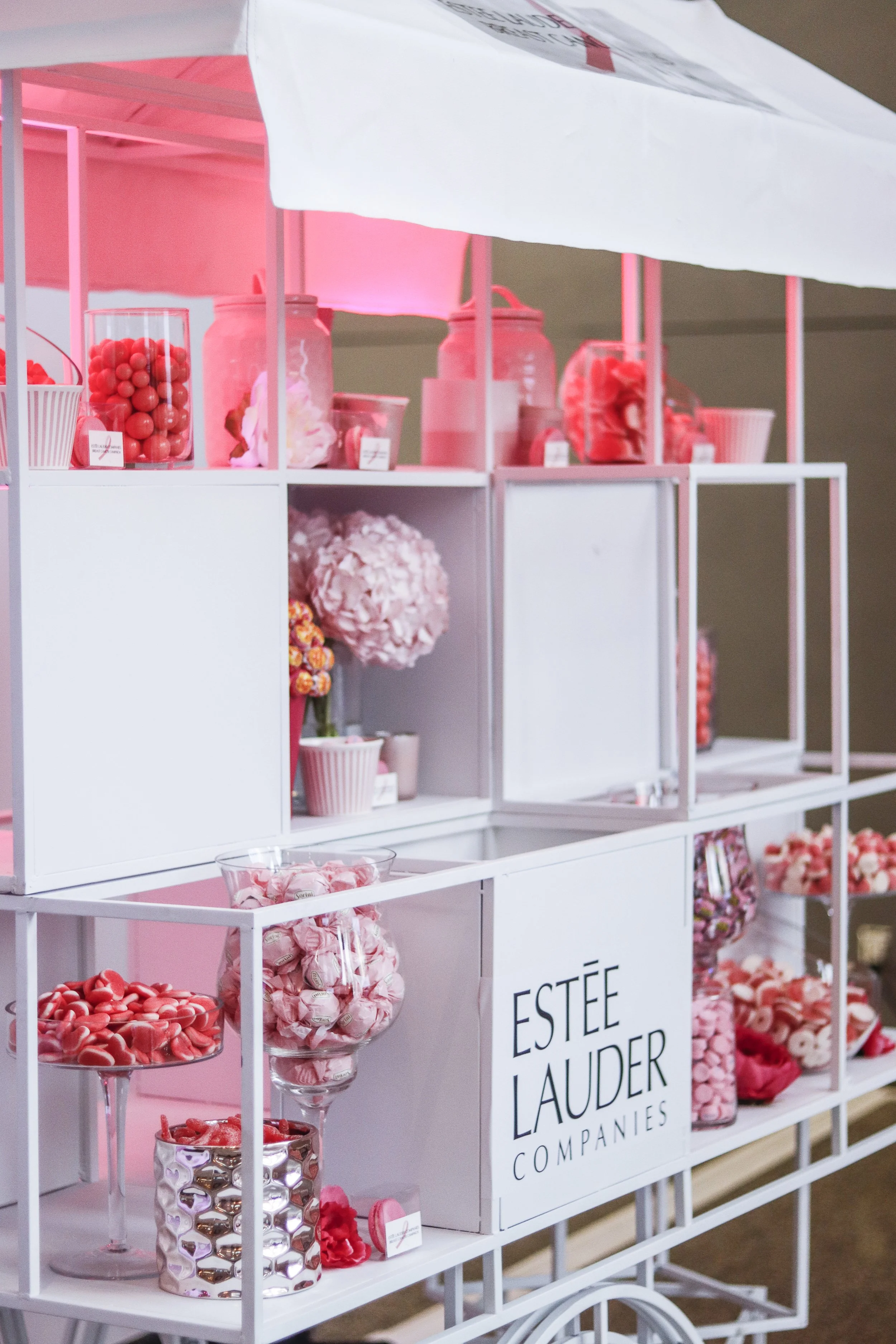 A display cart filled with pink and red candies and flowers, with a sign reading 'Estée Lauder Companies'.