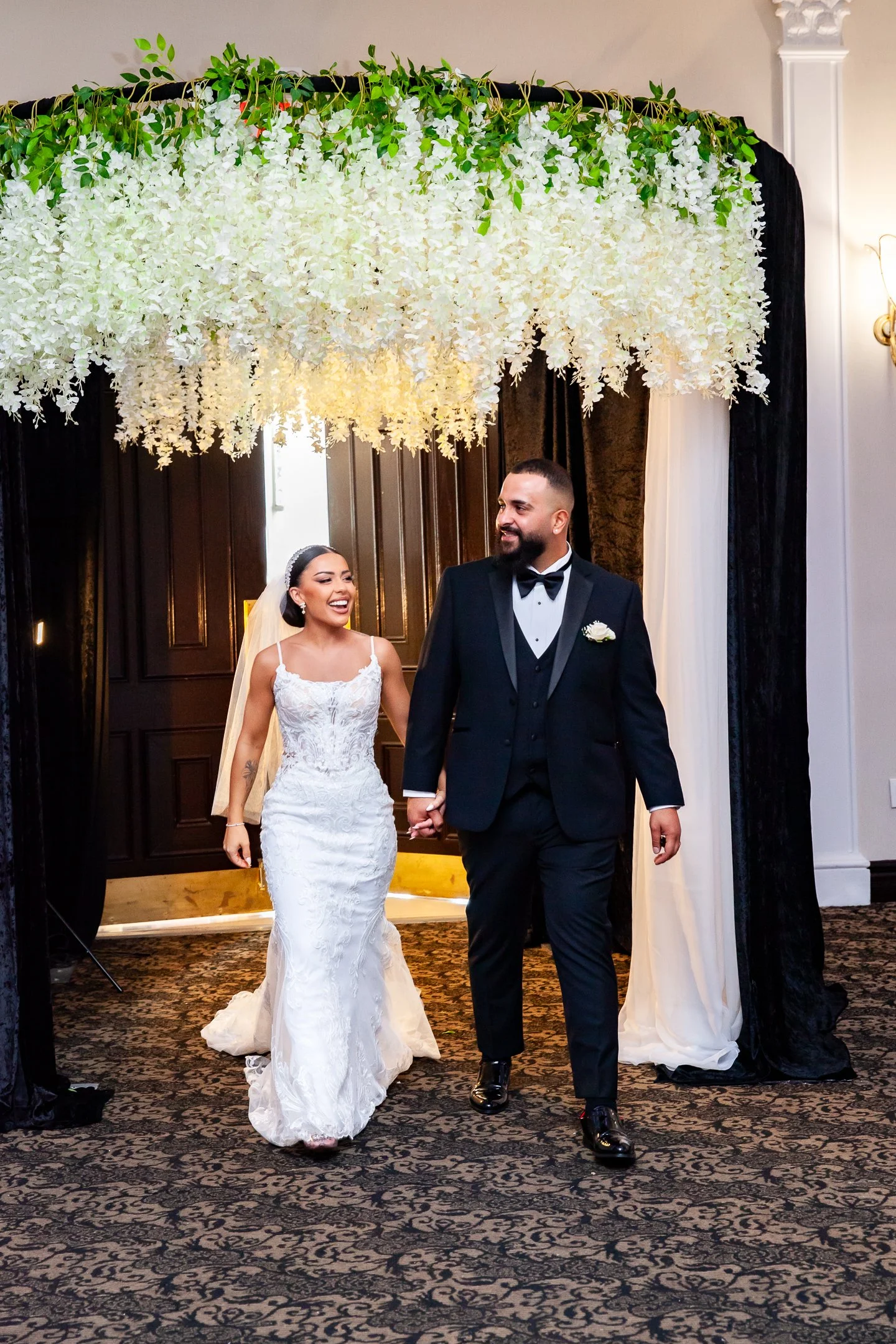A bride and groom walking hand in hand under a floral wedding arch, smiling and looking at each other.
