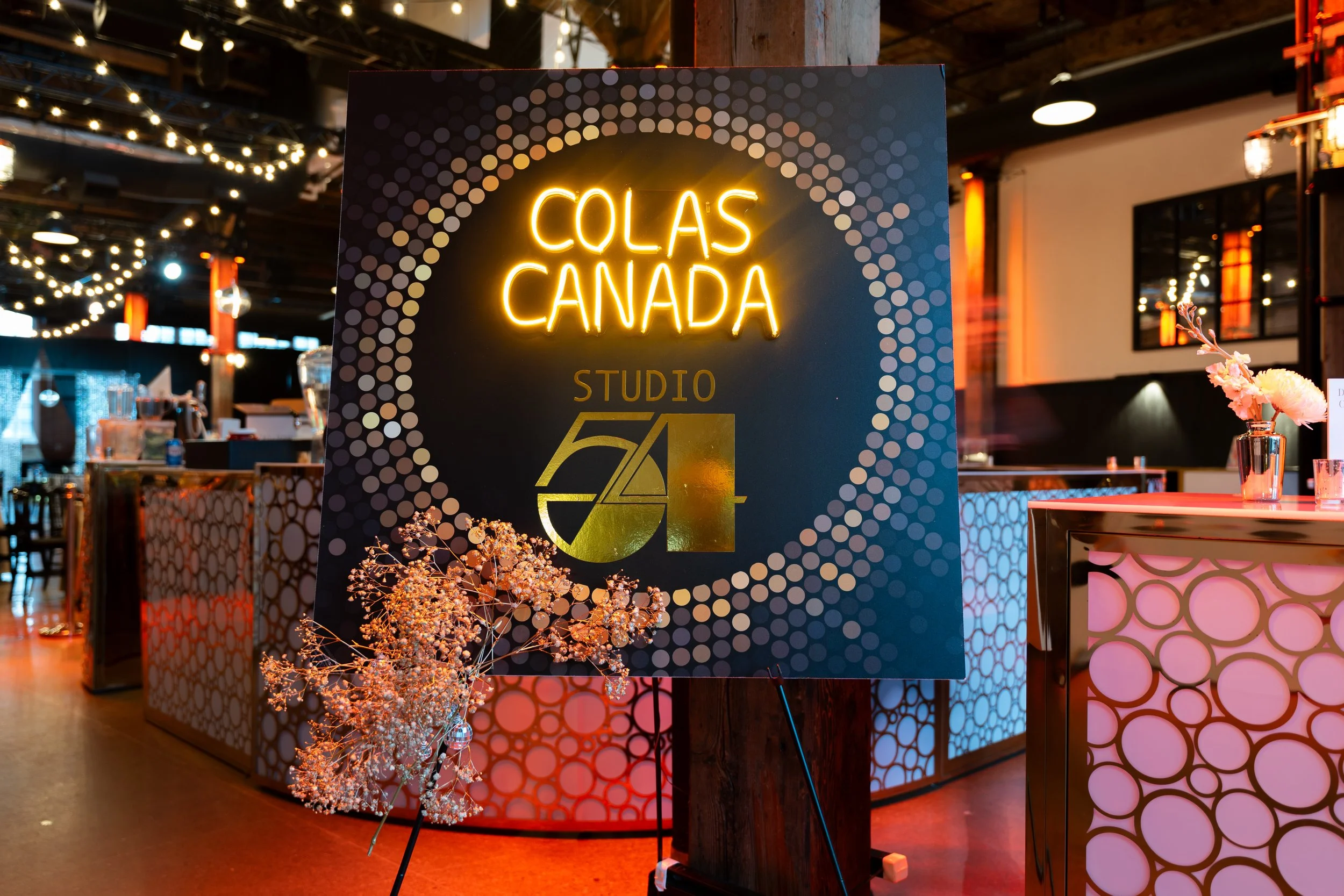 Neon sign reading 'COLAS CANADA' with a dot pattern background in a dimly lit indoor event space, decorated with flowers and illuminated string lights.