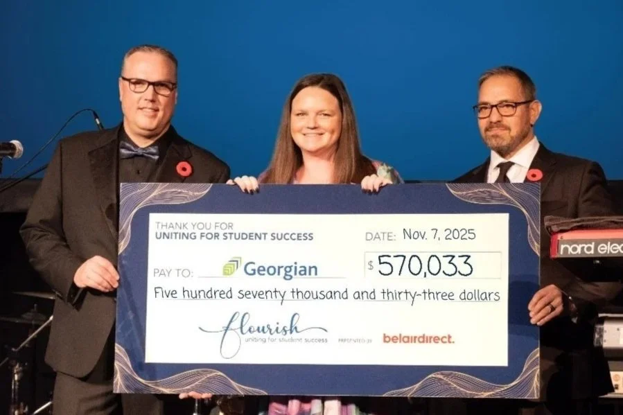 Three individuals standing together holding a large ceremonial check made out to Georgian for $570,033, dated November 7, 2025, at an event celebrating student success.