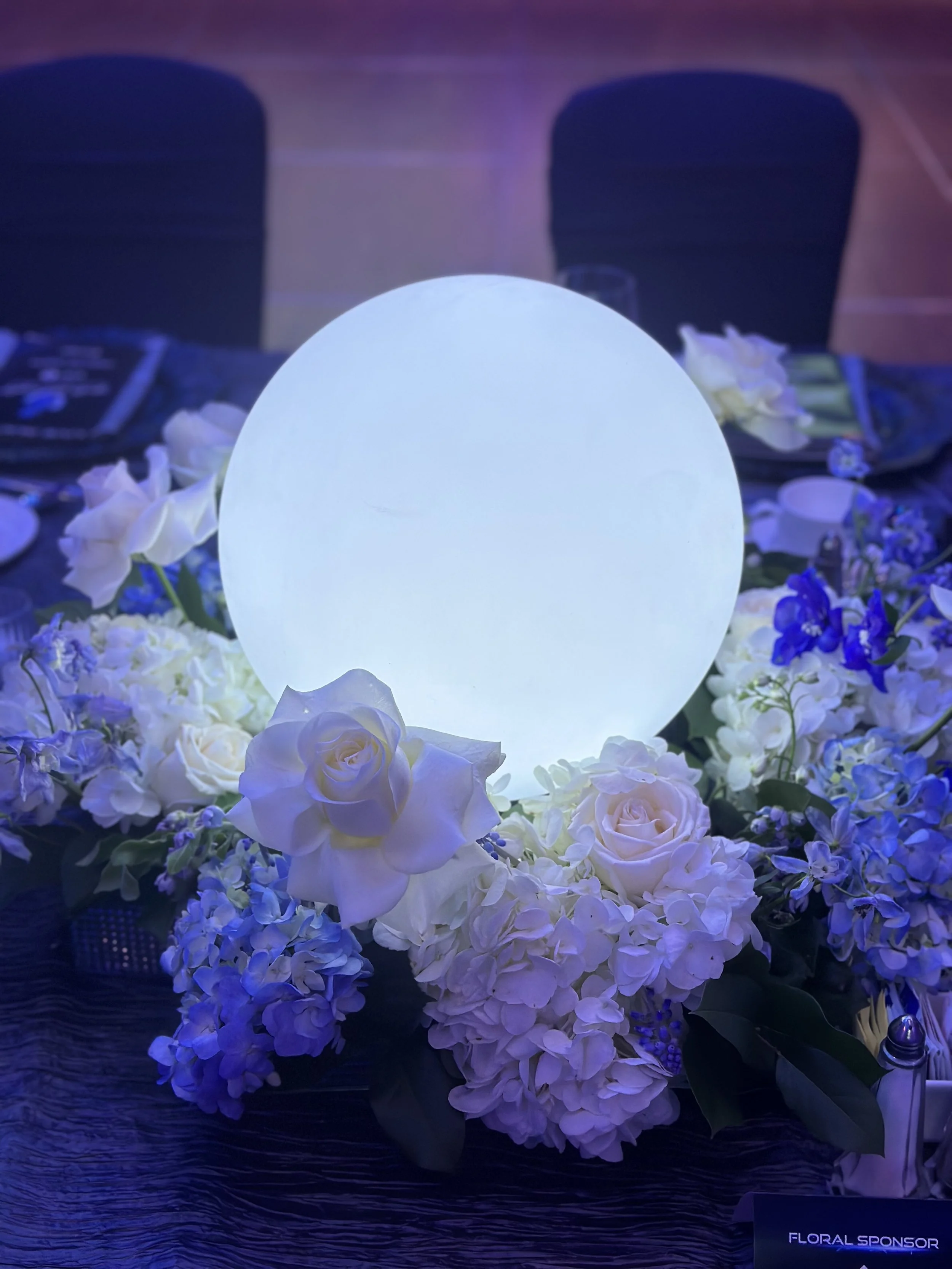 A romantic table setting with white roses, hydrangeas, and purple flowers, illuminated by a glowing orb, on a dark textured table, with chairs in the background.