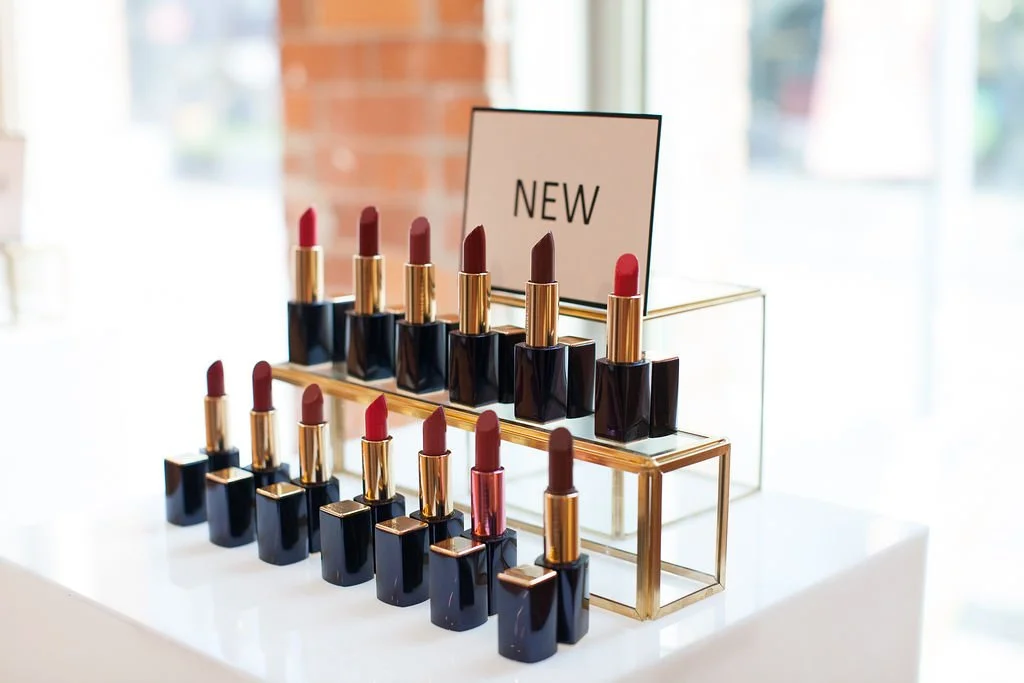 Display of various lipsticks on a white surface with a sign that says "NEW" in the background, inside a modern store or showroom.