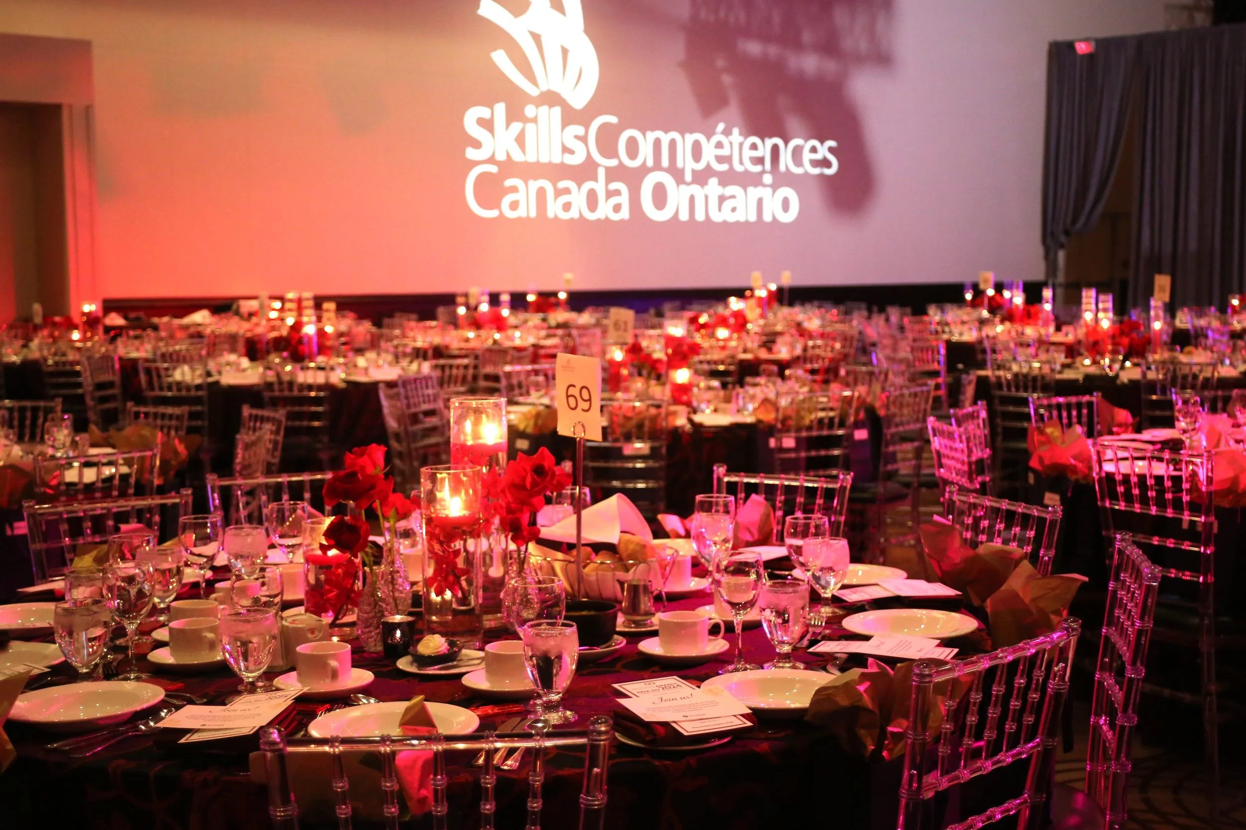 Event space with multiple round tables decorated with candles, flowers, and tableware, set for a formal dinner, featuring a large screen displaying the logo and text 'SkillsCompétences Canada Ontario'.