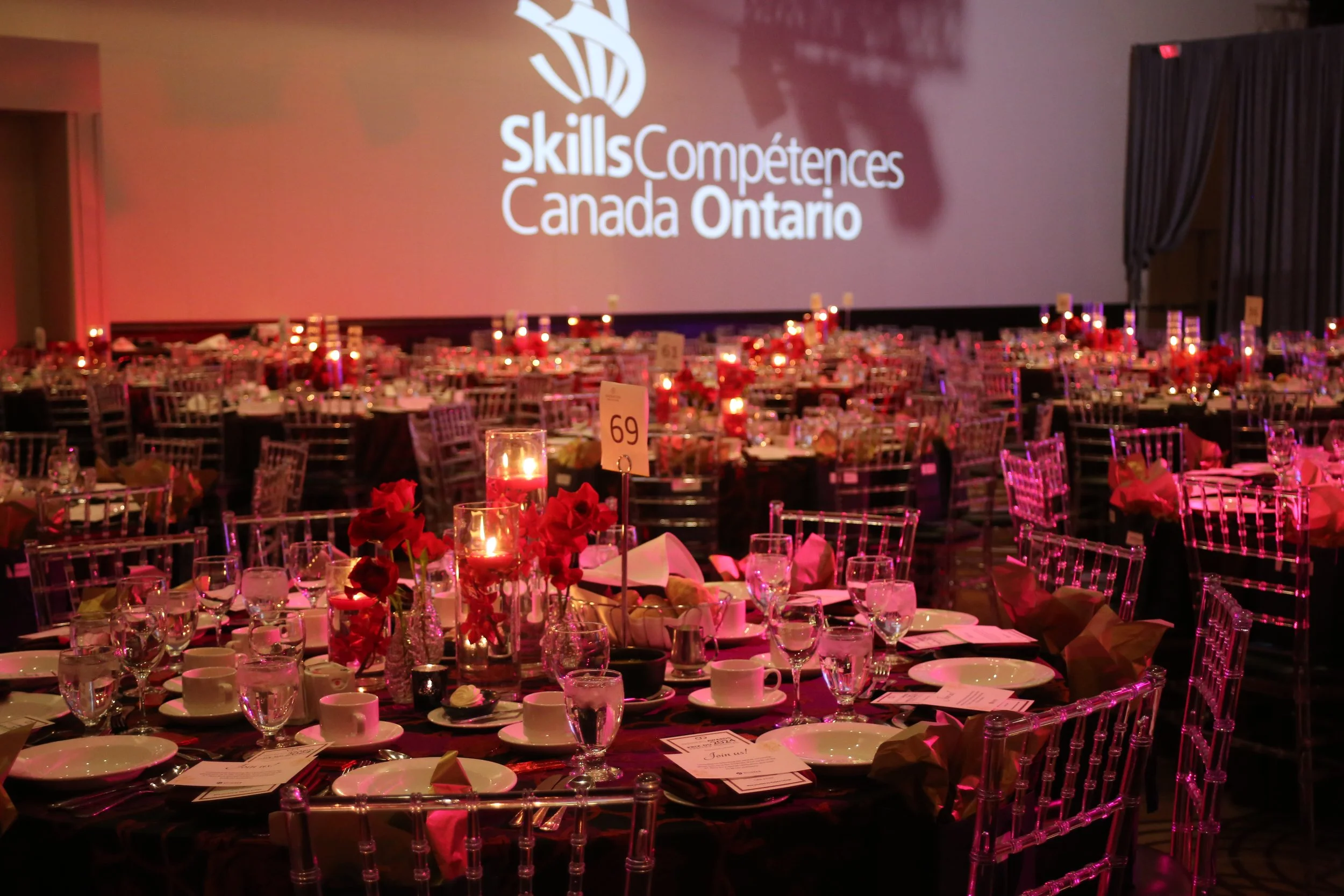 A large conference room decorated for an event with multiple tables set with glassware, plates, and candles. In the background, a large screen displays the logo and text for Skills Competences Canada Ontario. The room is dimly lit with pinkish-red li