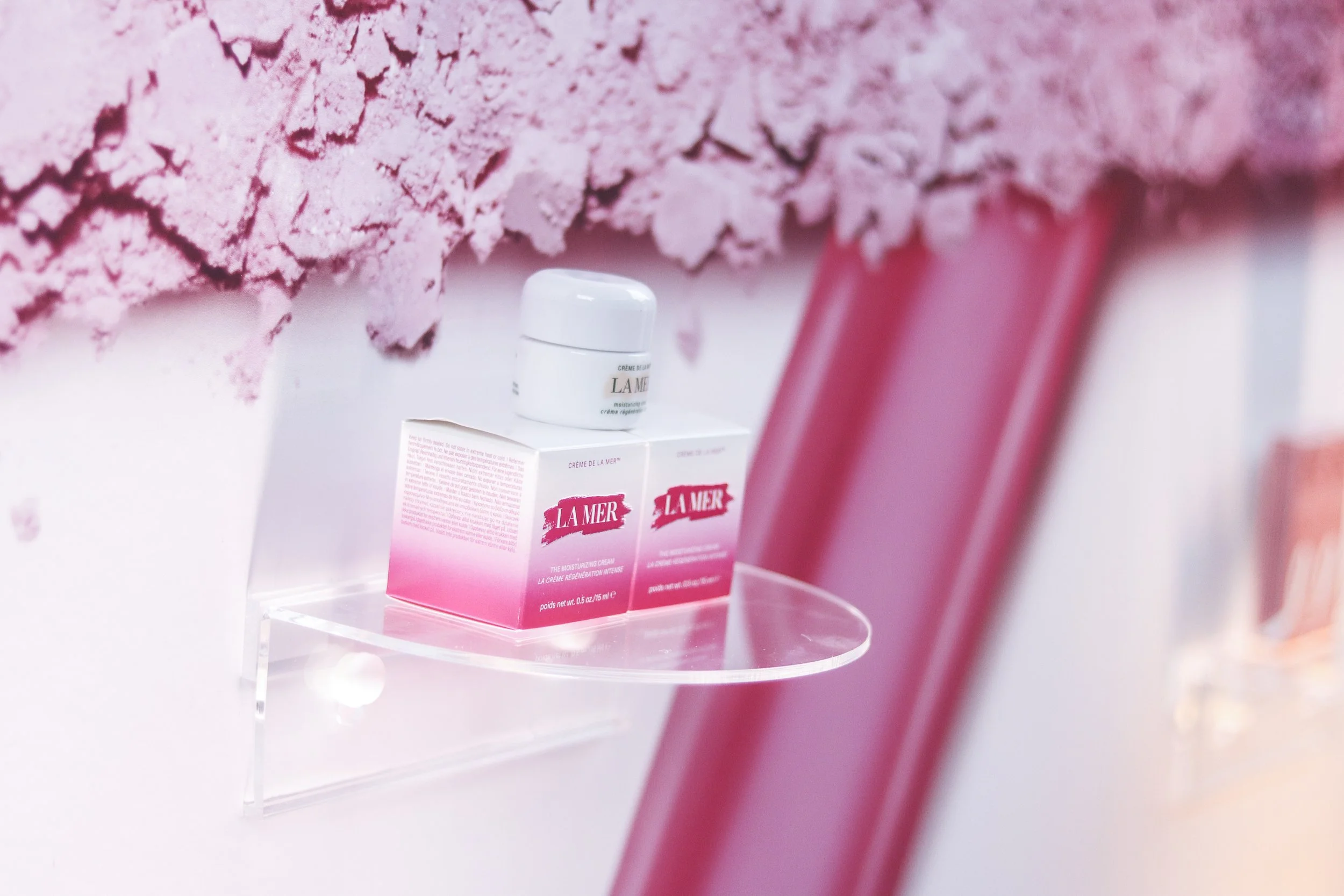 A small white jar of La Mer moisturizer product on a clear stand with pink packaging, placed in front of a textured pink wall with rough, irregular surface, and pink cylindrical objects to the side.