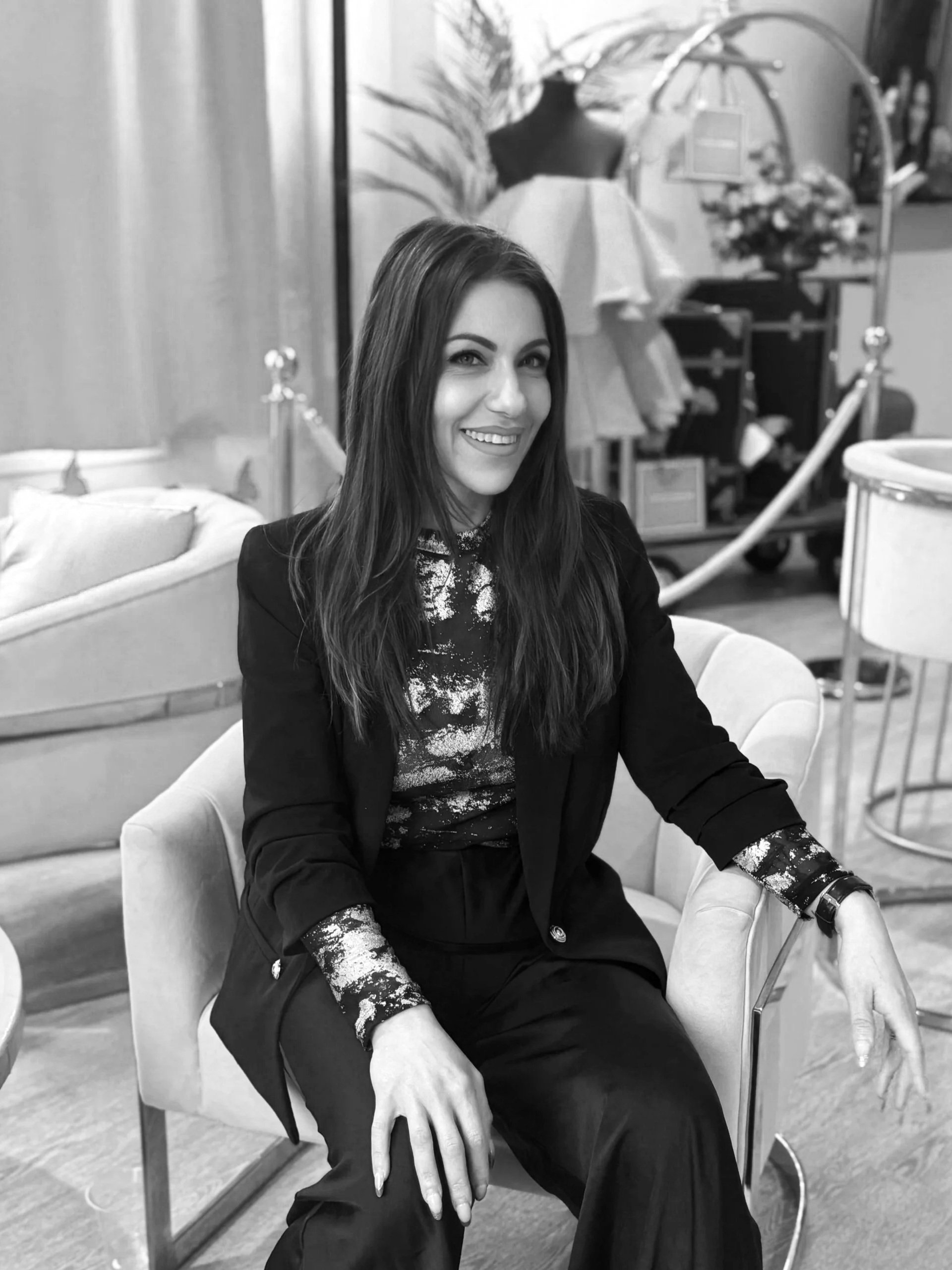 A woman sitting in a chair smiling at the camera, in a room with furniture and decorative items in the background.
