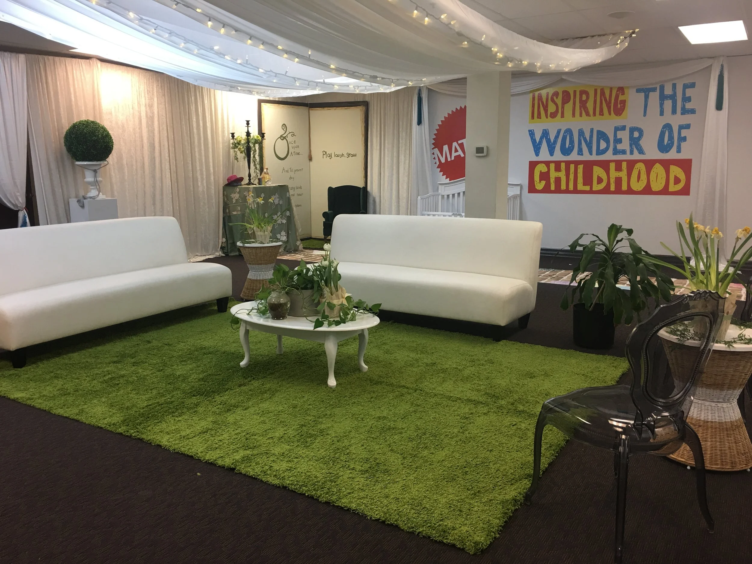 An indoor event space with white couches on a green rug, potted plants, decorative curtains, and a banner that reads inspiring the wonder of childhood, Mattel Head Office Designed by Alexandria Design House. Spring Press Preview 