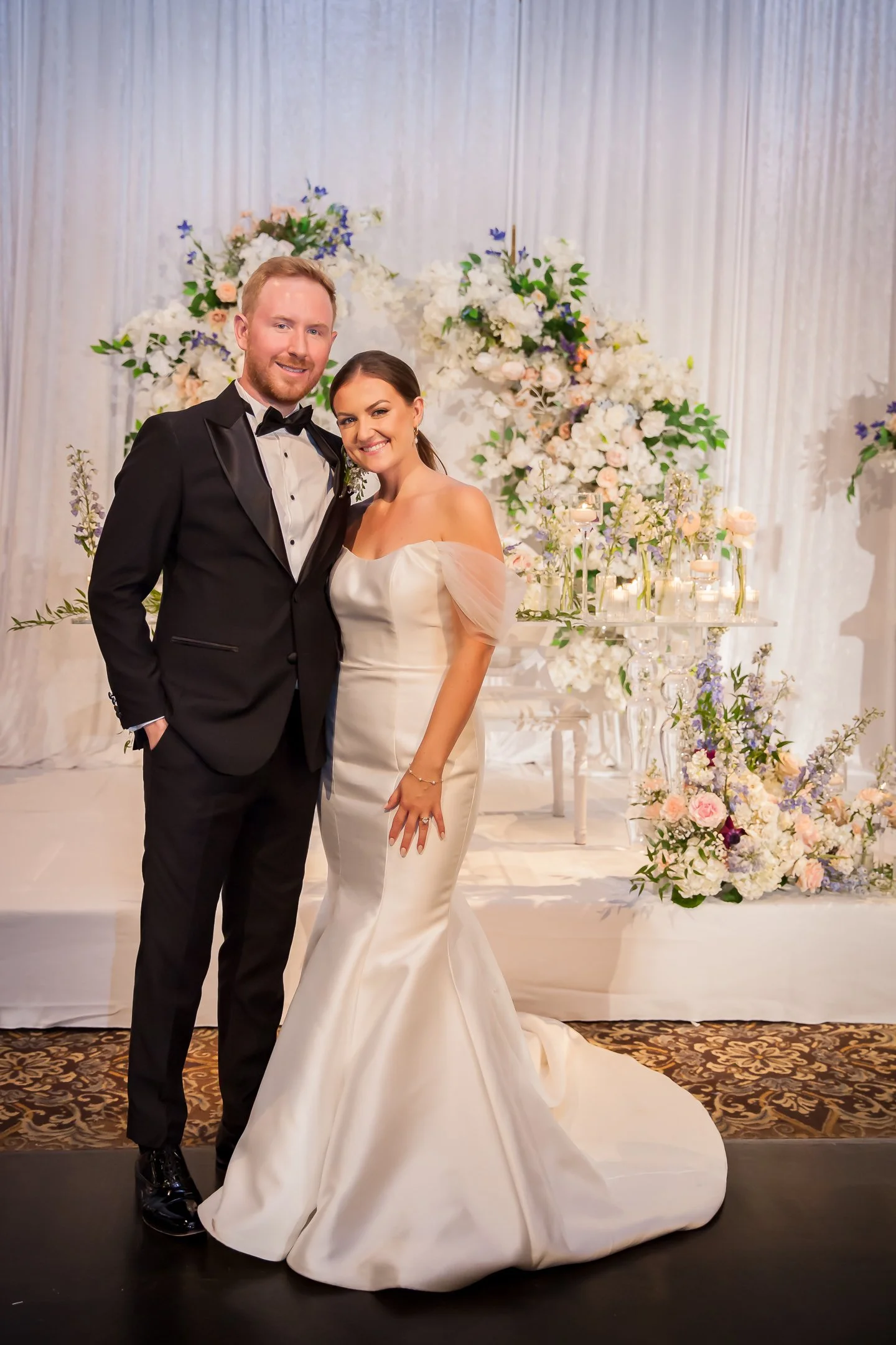 A bride and groom standing together at their wedding, with the bride in a white off-shoulder wedding gown and the groom in a black tuxedo, in front of a floral arrangement backdrop.