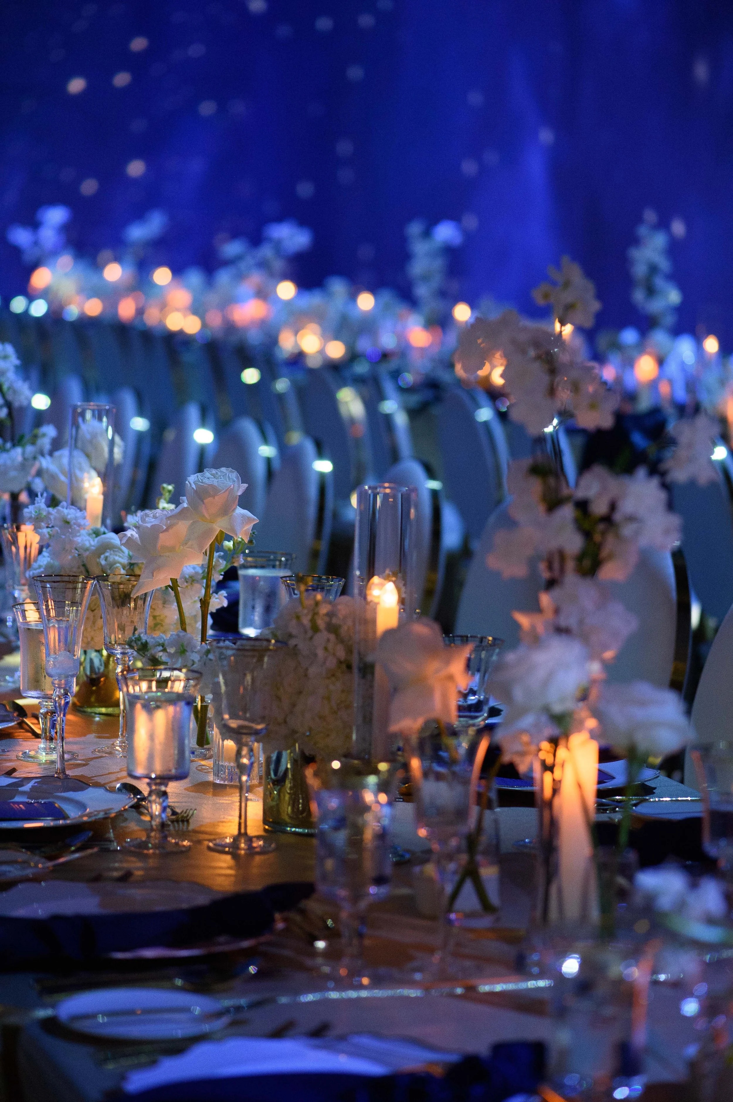 Elegant banquet table with white floral centerpieces, candlelit decor, and chairs set for an event in a dimly lit room with blue lighting.
