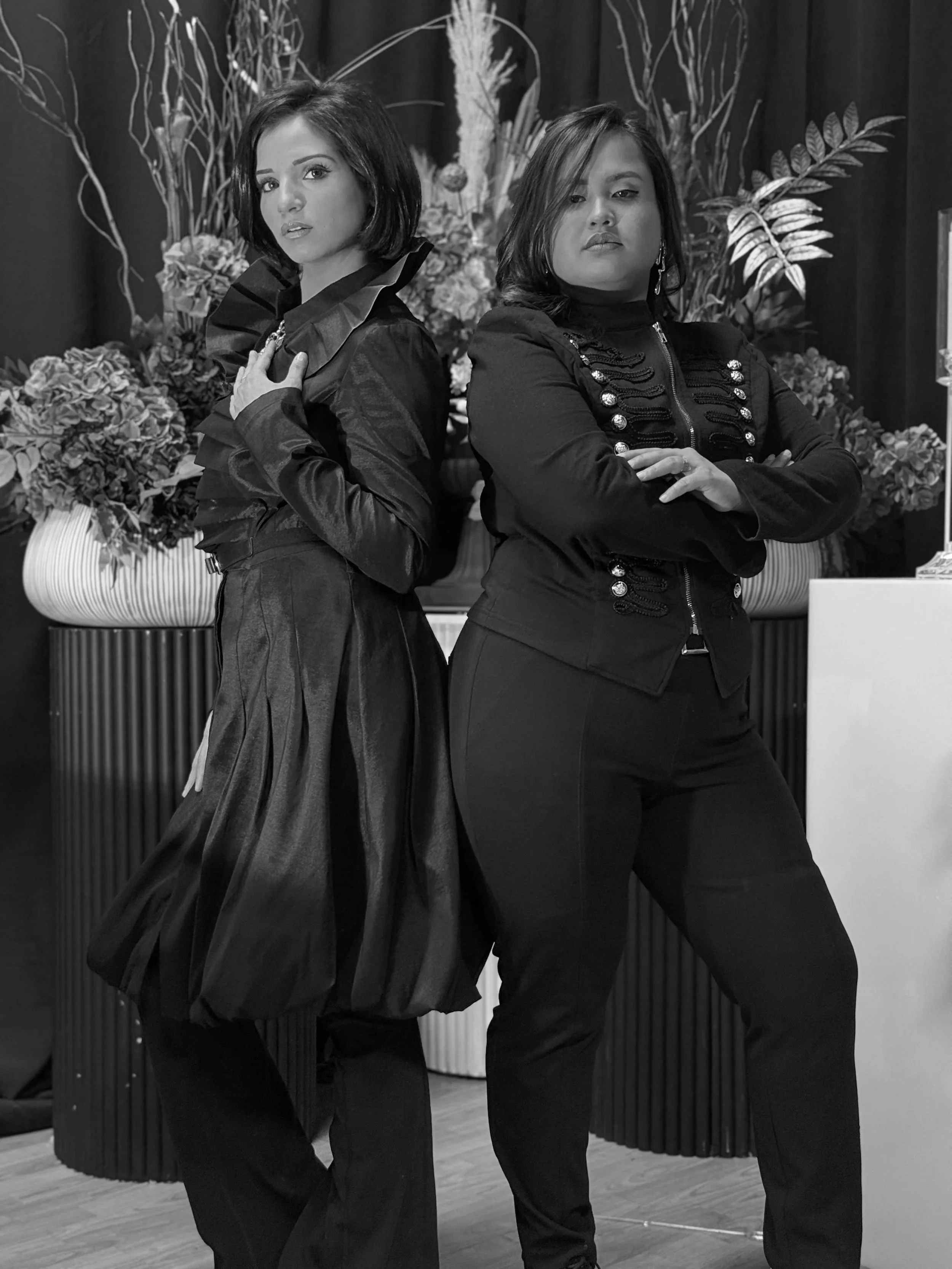 Two women in fashionable black outfits pose with arms crossed, in front of floral arrangements and plants, in a stylish indoor setting.