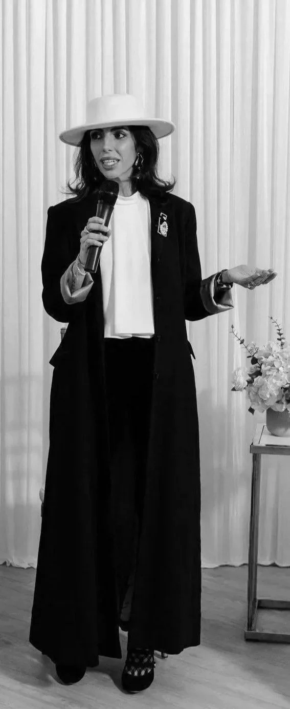 A woman dressed in a dark, long blazer and wide-legged pants, wearing a large wide-brimmed hat, holding a microphone and speaking, standing in front of light-colored curtains with a small table and flower arrangement nearby.