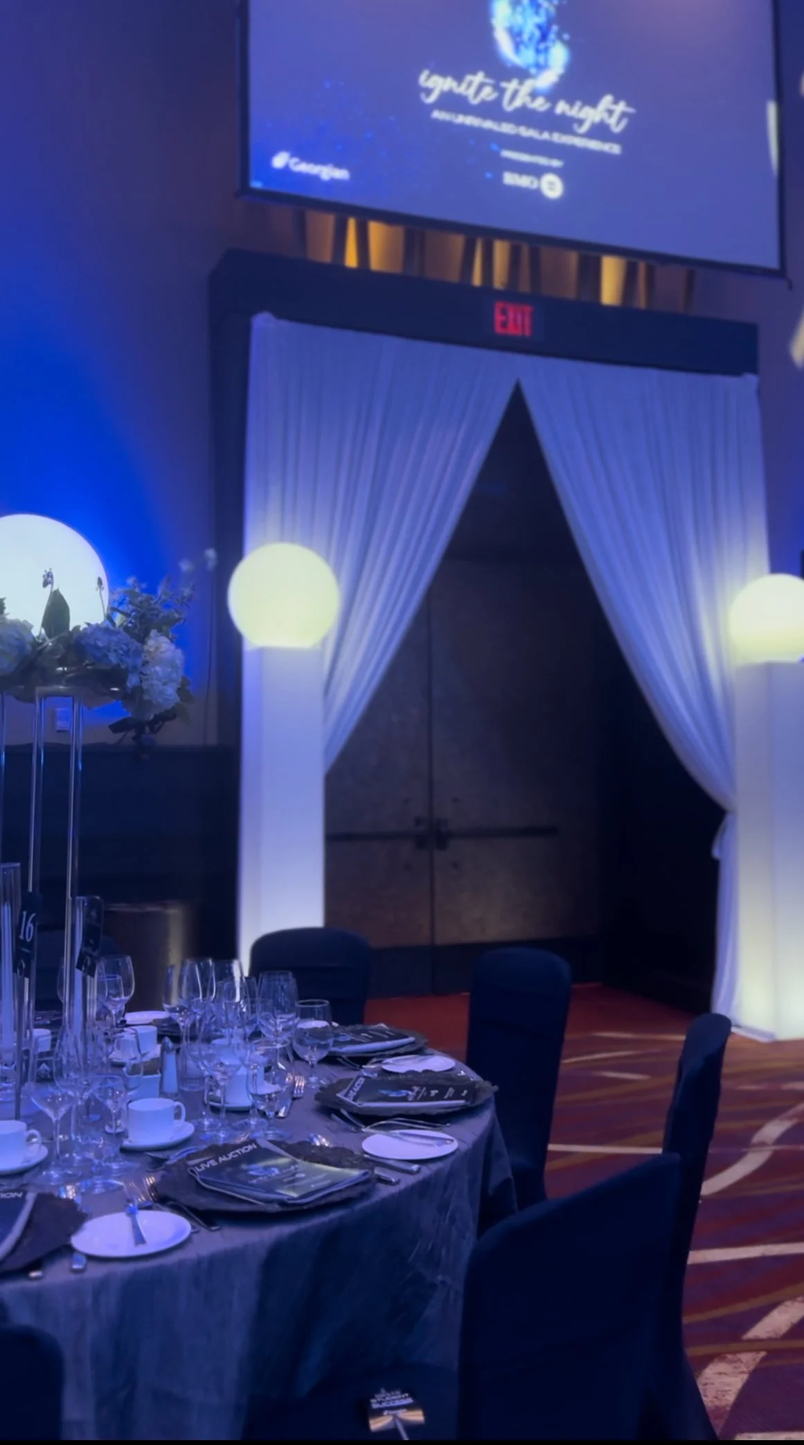 A decorated banquet table set for a formal event with black chairs, wine glasses, and floral centerpieces, in a dimly lit room with a blue and purple color scheme, and a white draped entrance or exit area with an illuminated sign and a large screen a