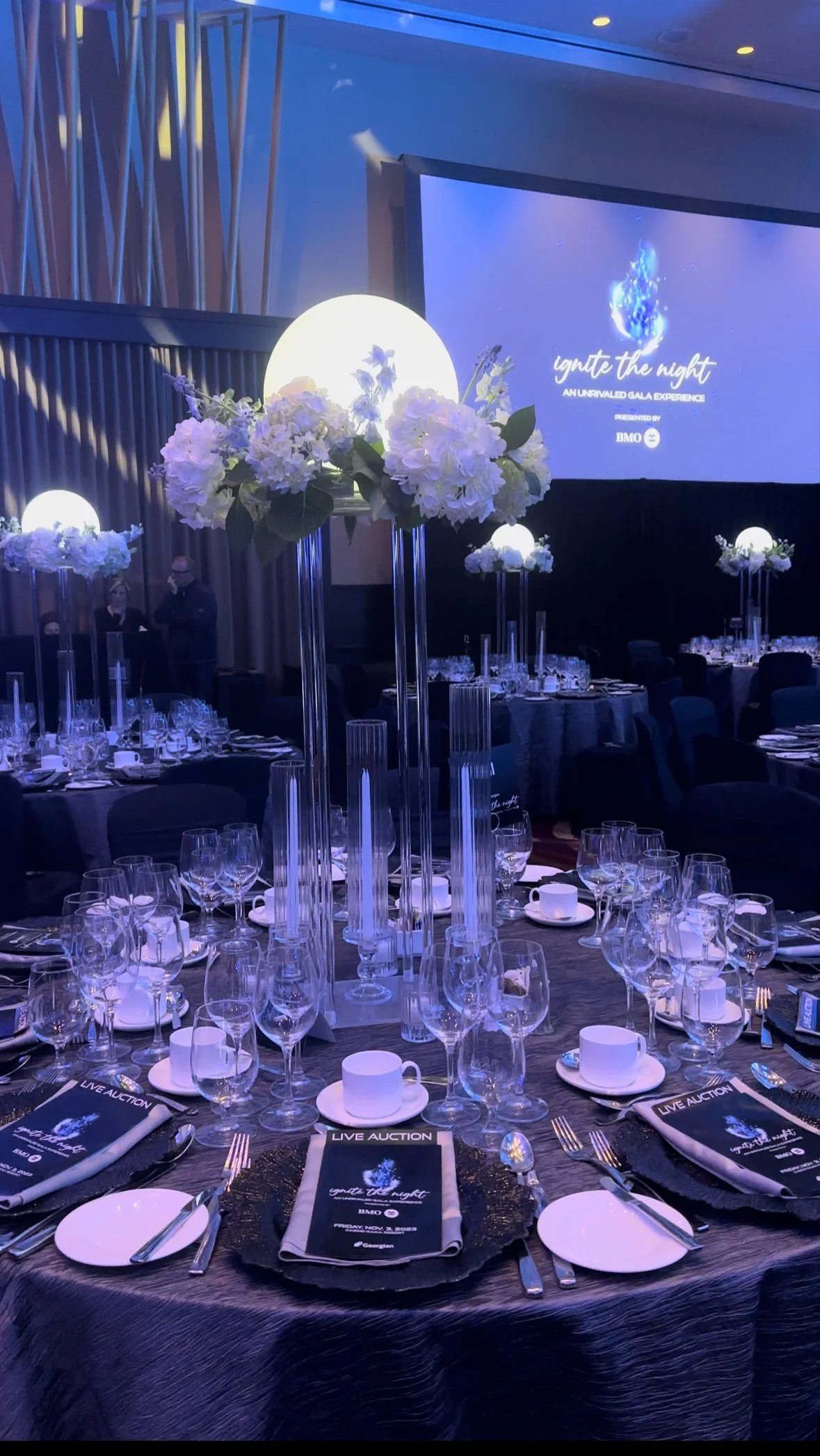 Elegant banquet table with tall floral centerpieces featuring white flowers and greenery, glassware, and place settings. A large screen in the background displays a logo and text about a gala event called "ignite the night" presented by BMO. The room