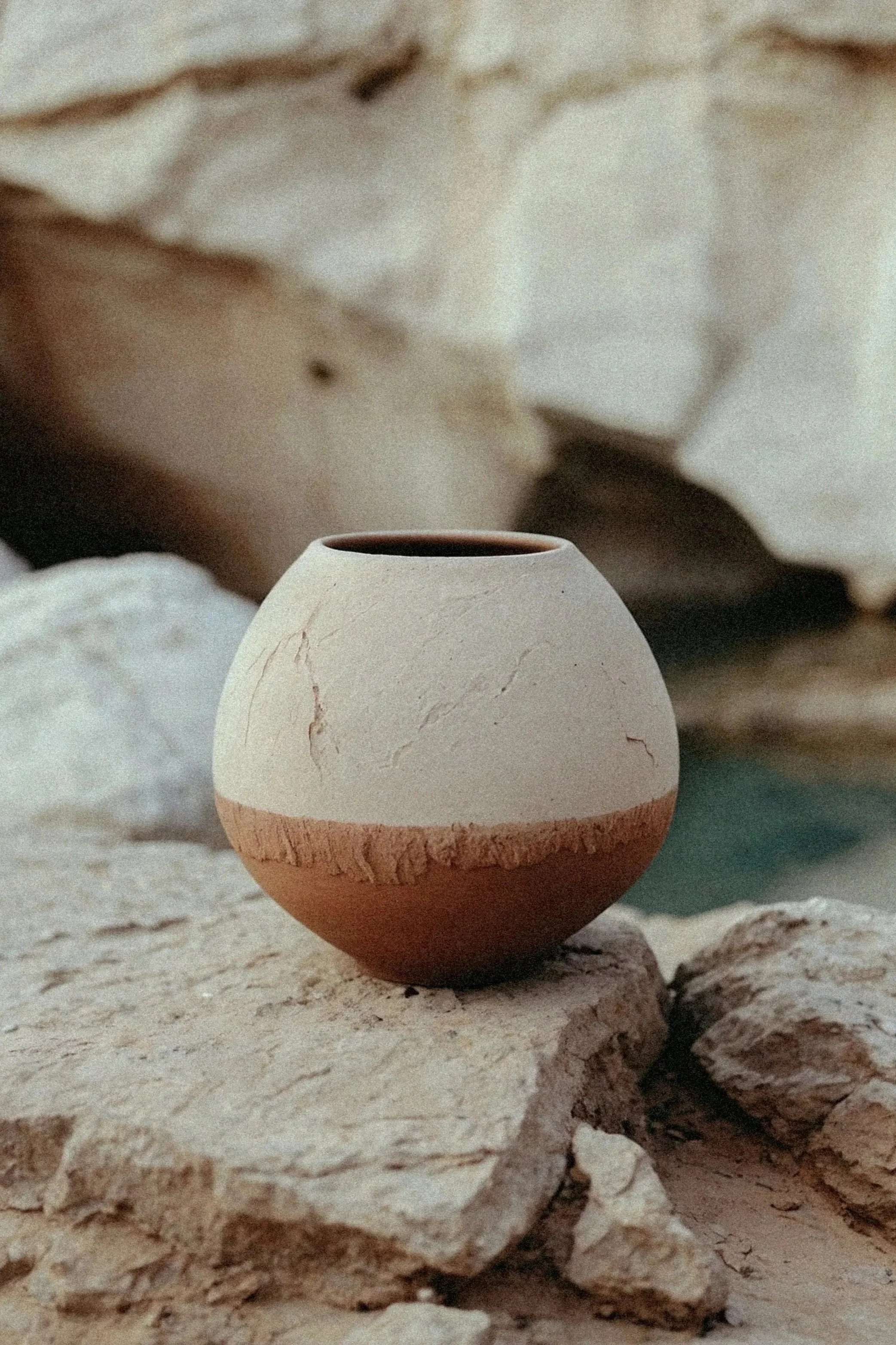 A two-tone clay pot with a cream upper half and a reddish-brown lower half is placed among rocks and stones.