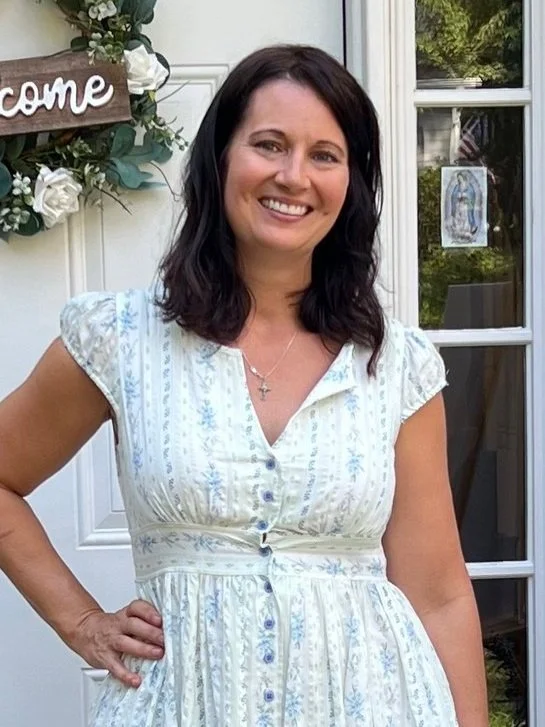 A smiling woman with dark hair wearing a light-colored, short-sleeved, button-down dress with a floral pattern, standing outside near a white door decorated with a floral wreath and a welcome sign.