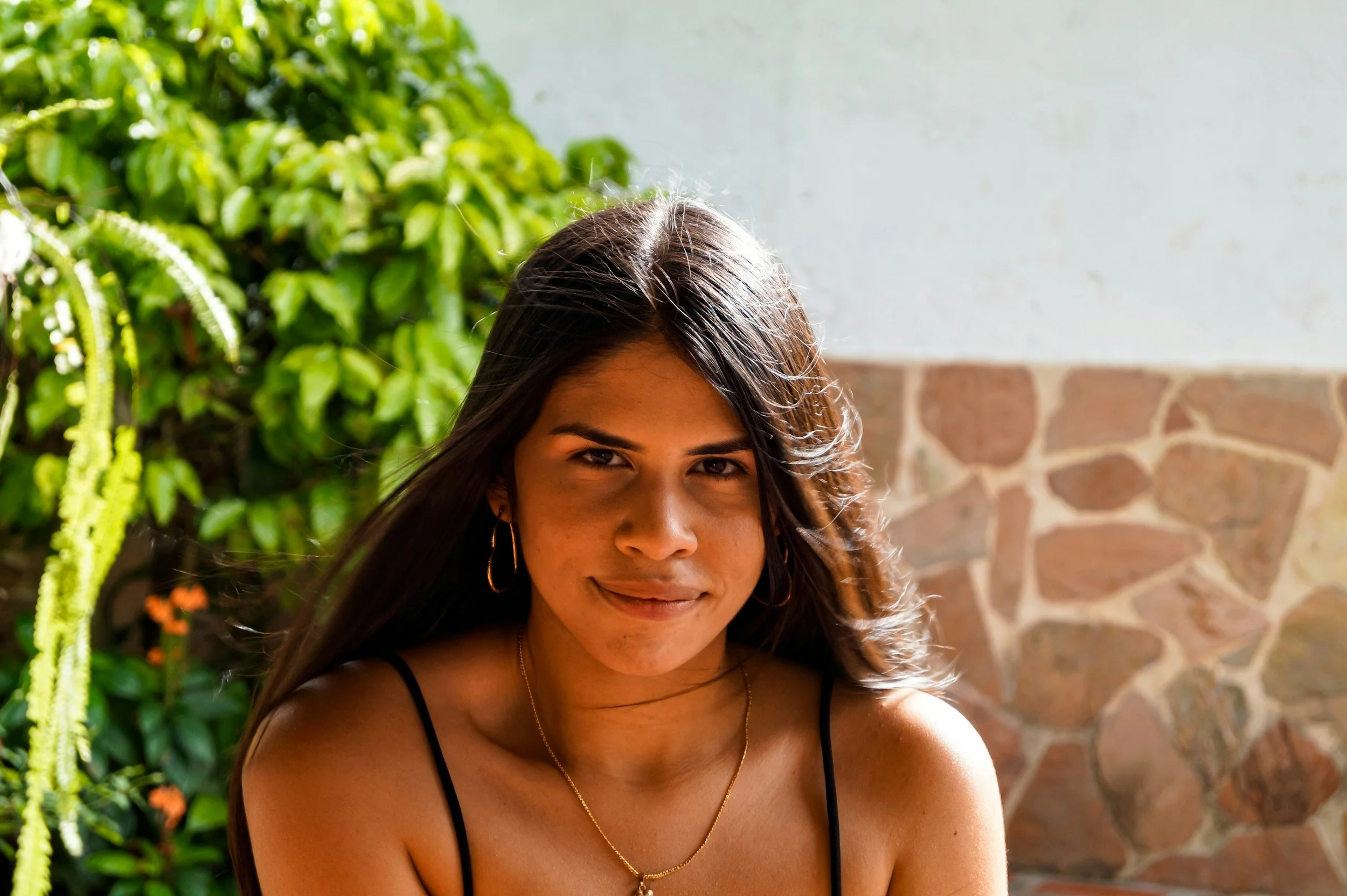 A woman smiling outdoors with green plants and a stone wall in the background.