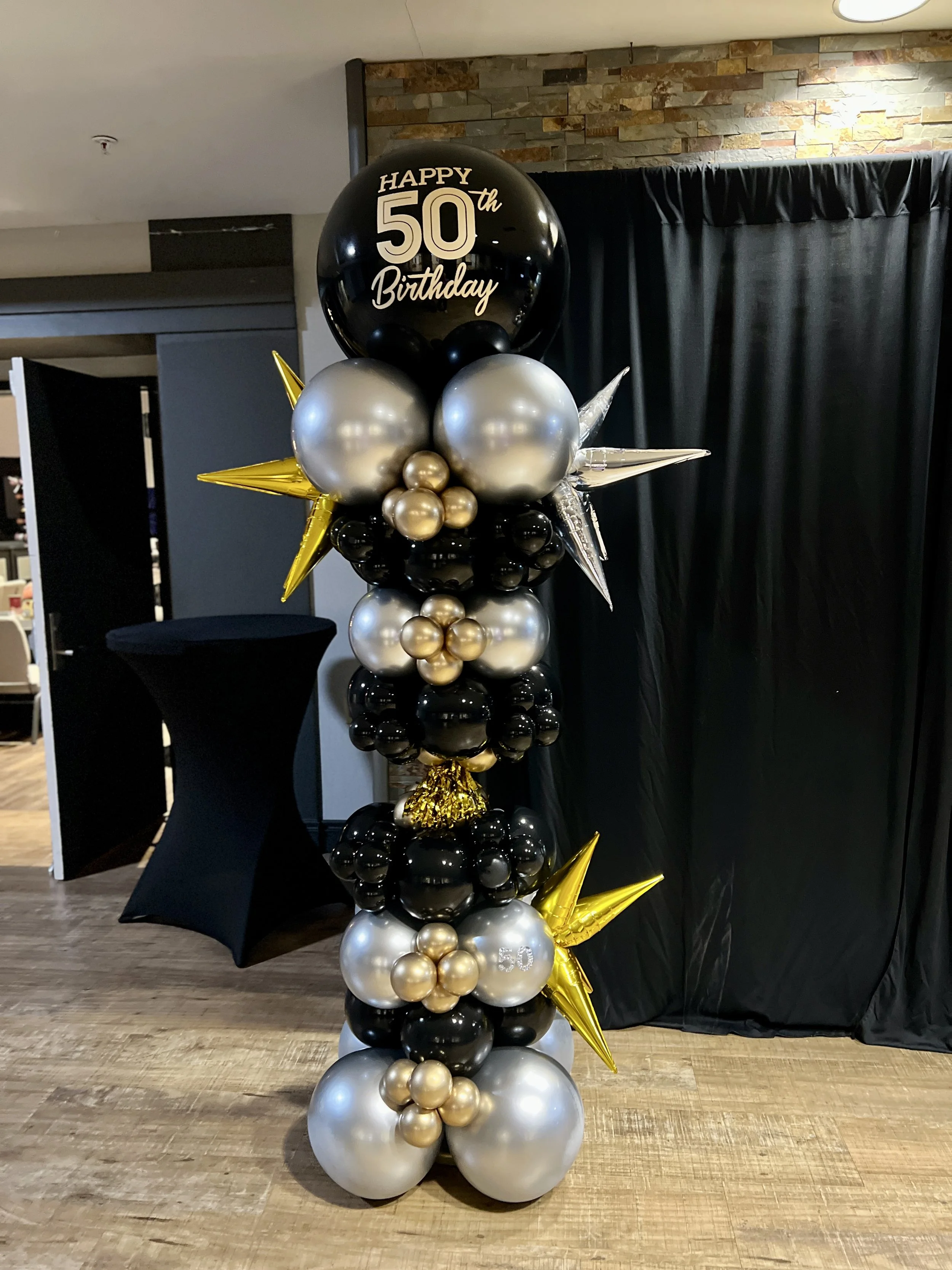 Balloon column with black, gold, and silver balloons celebrating a 50th birthday, topped with a large black balloon reading "Happy 50th Birthday" and decorated with gold stars.