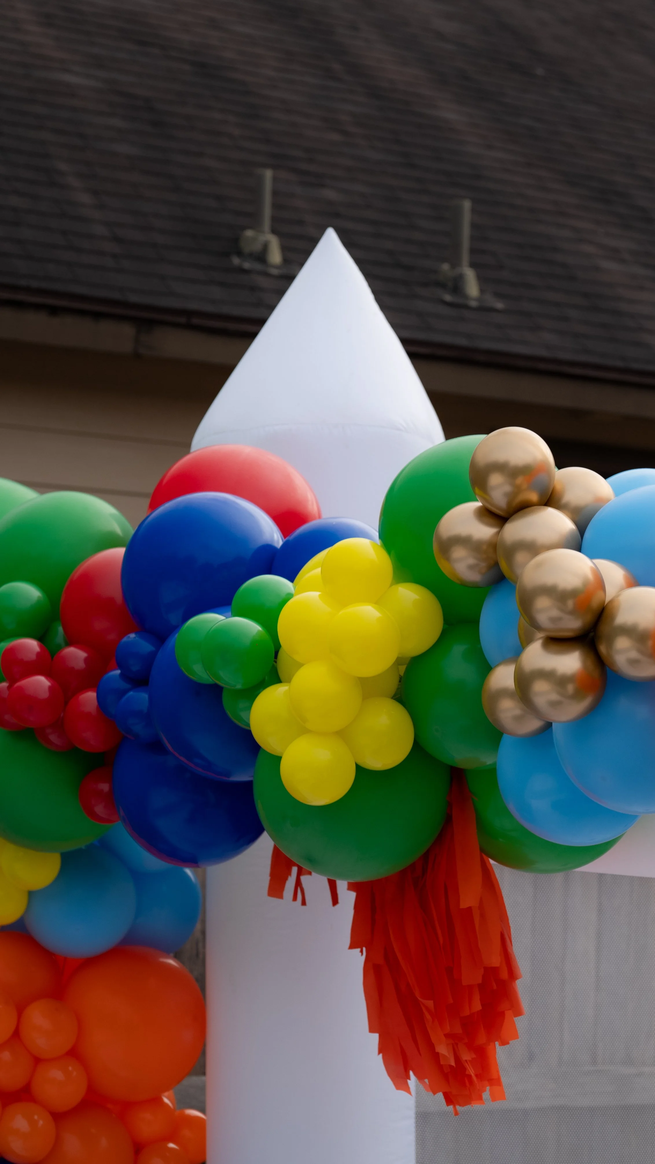 Colorful balloons decorated on a white structure with an orange fringe, set against a building with a shingled roof.